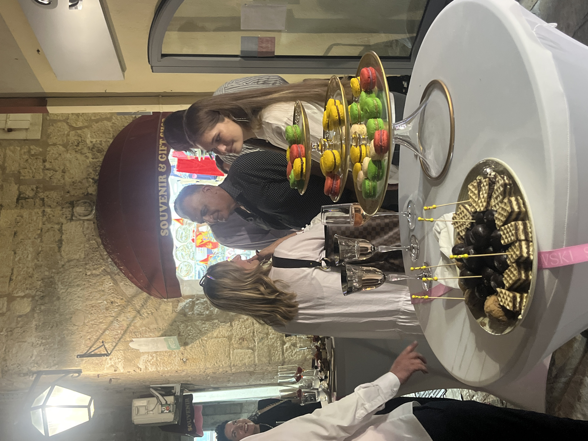 This media file captures an indoor event, likely a reception or party, with a group of people gathered around a table laden with desserts. The scene is set against a backdrop of stone walls, reminiscent of a historic building or a venue with rustic architecture.
The subjects in the image include several individuals, some actively engaged in conversation or observing the food. A woman with long brown hair is leaning in, looking at a display of colorful macarons. Next to her, a man in a patterned shirt is also visible. Another woman, wearing a white dress, stands further down the table. In the foreground, a man in a white shirt and black pants is partially visible, extending his hand.
The objects on display are primarily food items. There are tiered platters filled with brightly colored macarons in shades of red, yellow, and green. Another platter holds chocolate-covered items and wafer cookies, artfully arranged. Several champagne flutes and wine glasses are also present on the table, suggesting a celebratory occasion.
The lighting appears to be artificial, indicating an indoor event, possibly in the evening or a dimly lit space. The overall atmosphere seems social and relaxed, with people enjoying the food and company.
Visible text includes "SOUVENIR & GIFT SHOP" on a sign above a display window in the background. Another partial text "VSKI S" is visible on a pink ribbon tied around the table. The context provided places the location as Budva, Montenegro.