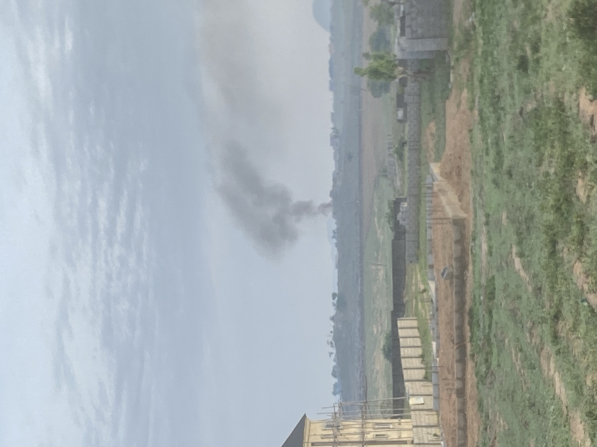 The image shows a hazy, overcast sky over a partially constructed area near Lugbe, Nigeria. A plume of dark smoke rises in the distance. The ground is mostly undeveloped with areas of green vegetation and exposed dirt. There are several partially-built brick structures visible, some with scaffolding.
