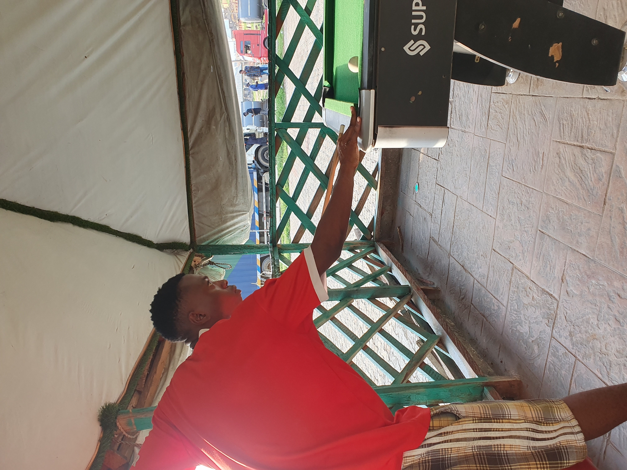 In Lokoja, Nigeria, under a white tarp roof supported by bamboo poles, a young Black man in a red t-shirt and plaid shorts is playing pool. He is positioned on the right side of the frame, leaning over the green pool table, holding a cue stick and aiming at a white ball. A single white ball rests on the table. The pool table itself is black with the brand name "SUPER" visible. Behind the man, a green wooden lattice fence separates the playing area from an outdoor tiled patio. Beyond the fence, trucks and people are visible, suggesting the location is in a public area or marketplace. The lighting suggests it is daytime.
