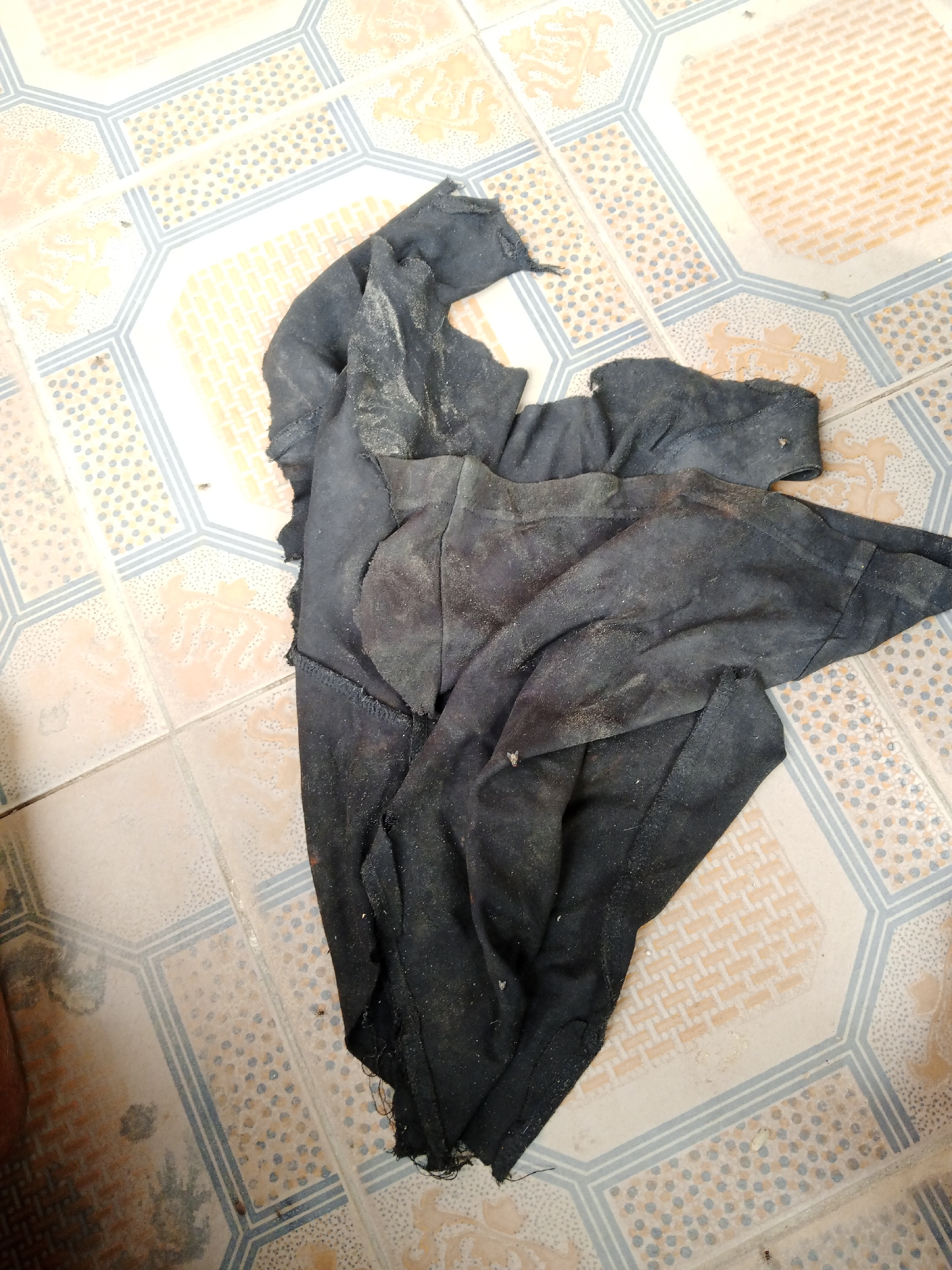 A pile of dirty, black fabric lies crumpled on a tiled floor. The fabric appears to be made of a rough material and is covered in a fine, light-colored dust or dirt. The tiles are patterned with alternating beige and white octagons, with blue and grey borders. There are also some smaller orange and cream colored tiles visible. The scene is lit from above. The overall impression is one of neglect or abandonment. There are no people or animals visible in the image, and no other objects besides the fabric and the tiles.