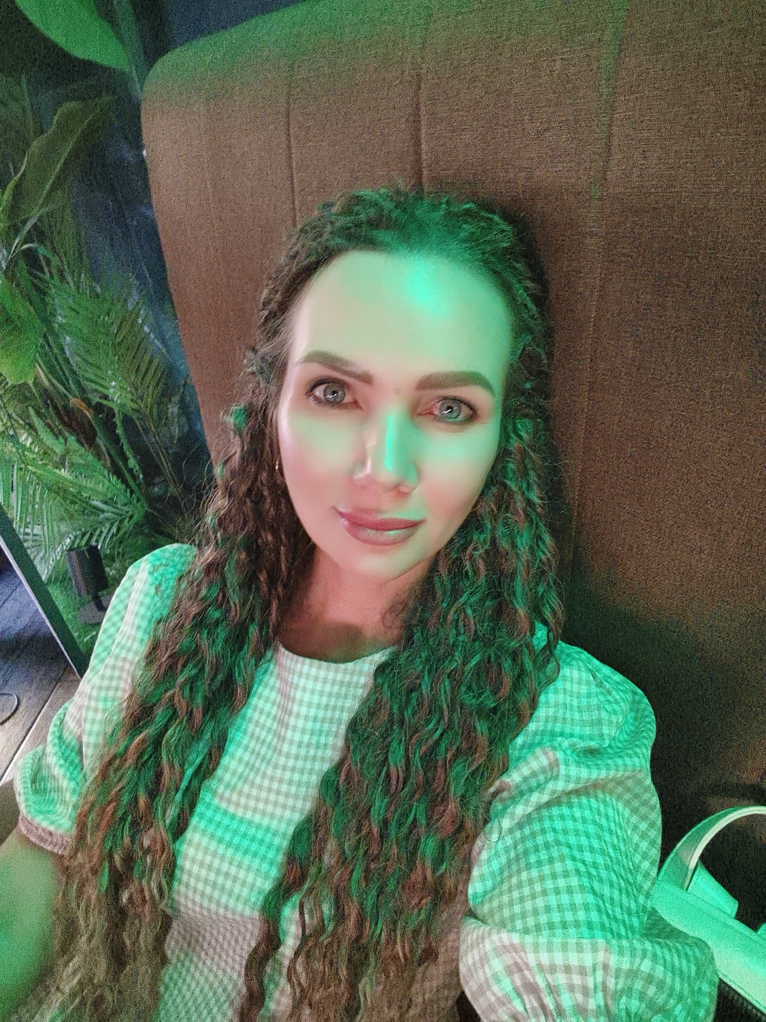A woman with long, curly brown hair and blue eyes is seated in an armchair. She is wearing a light-colored, checkered dress. The lighting in the scene is predominantly green, casting a neon glow on her face and clothing. To her left, there are lush green plants, some of which appear to be tropical foliage. The armchair is upholstered in a brown fabric, and the background is dark. The overall impression is that of an indoor setting, possibly a lounge or a dimly lit room with decorative plants. The image is a selfie, taken from a slightly high angle.