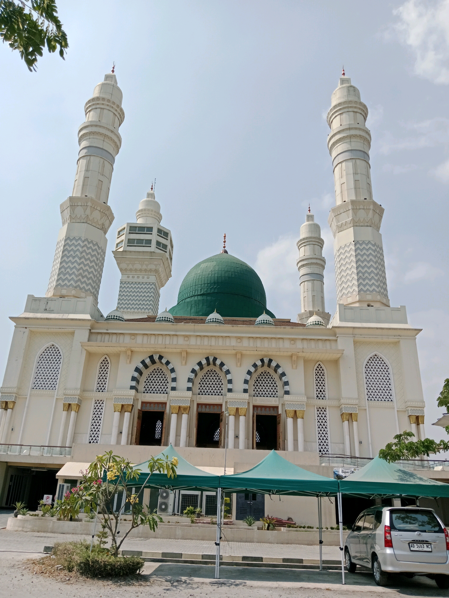 An exterior, low-angle, full shot captures a mosque with white and light grey facade under a bright, mostly clear sky with some scattered clouds. The mosque features a prominent green dome, four tall, slender minarets with decorative bands, and multiple arched windows with intricate white lattice work. The facade also includes gold-colored columns and decorative blue and black stripes above the entrances. In the foreground, a silver minivan is parked to the right, and two green canopies are set up in front of the mosque. A small tree with pink and yellow flowers stands in front of the canopies. The overall environment appears to be an urban setting in Karanganyar, Indonesia, with paved ground and some sparse vegetation. There are no people visible in the frame.