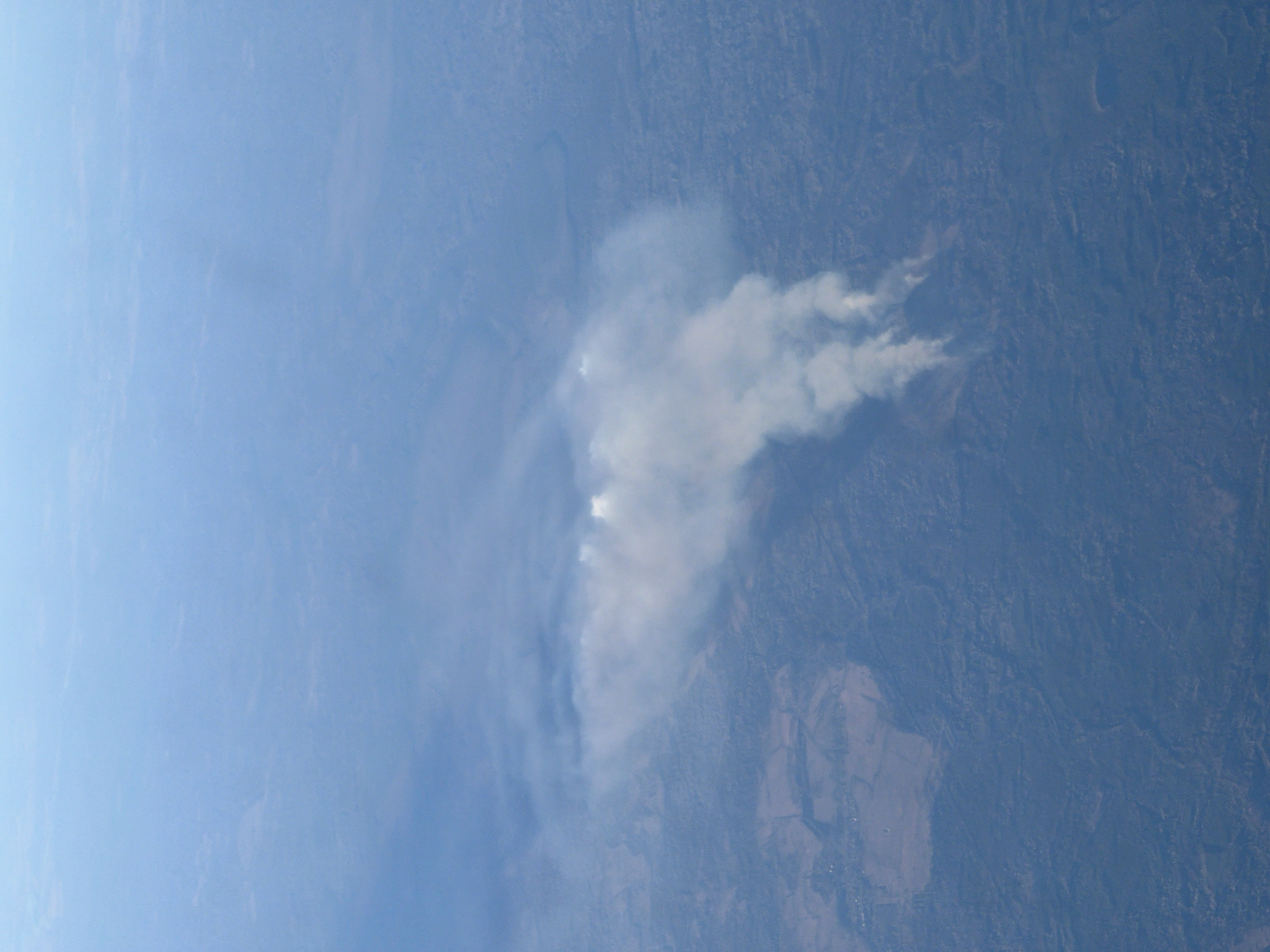 An aerial photograph displays a plume of white smoke drifting over what appears to be a rural landscape. The background shows terrain with varying textures and tones, possibly indicative of fields, forests, or natural land formations. The sky, heavily obscured with haze or smog, adds to the indistinct nature of the scene. This image does not appear to be located in the city of Saint Petersburg, Russia.