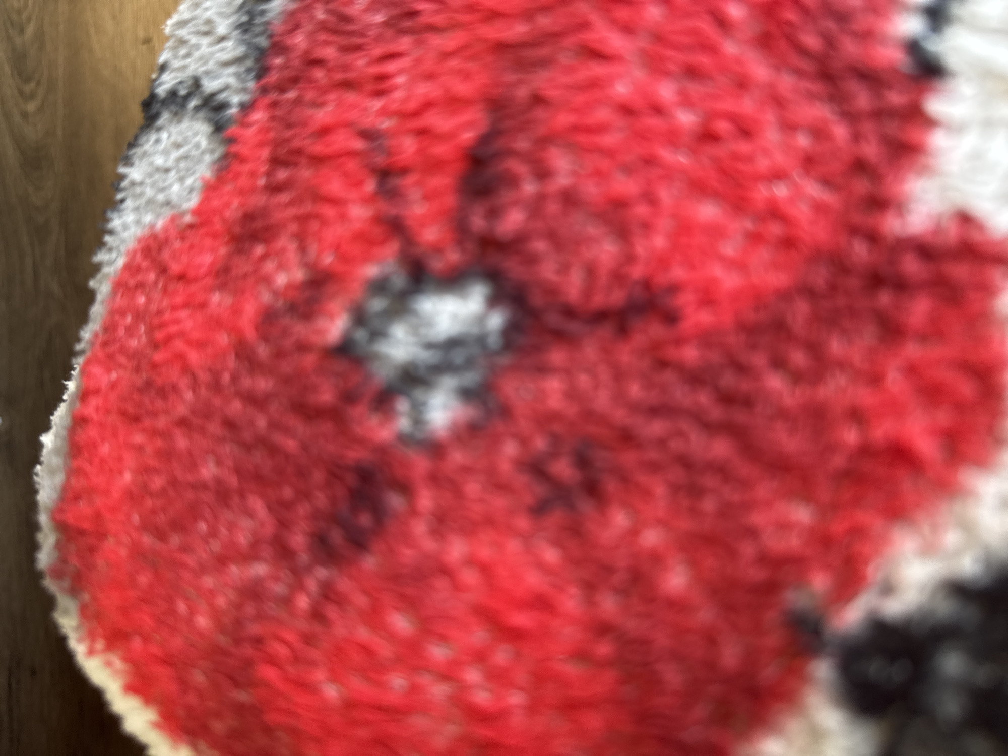 The image shows a close-up, blurred view of a fluffy, red, white, and black patterned textile, possibly a rug or blanket. The pattern appears to be abstract or depict a stylized image, with large red areas and interspersed white and dark grey/black patches. The texture is clearly visible due to the softness and fuzziness of the material. There are no people or specific identifiable objects present in the immediate foreground. The background is dark and out of focus, with a hint of wood grain suggesting a floor or furniture. The location is the city of Ridabu, Norway, but no specific landmarks or environmental details relevant to that location are discernible from the image itself.