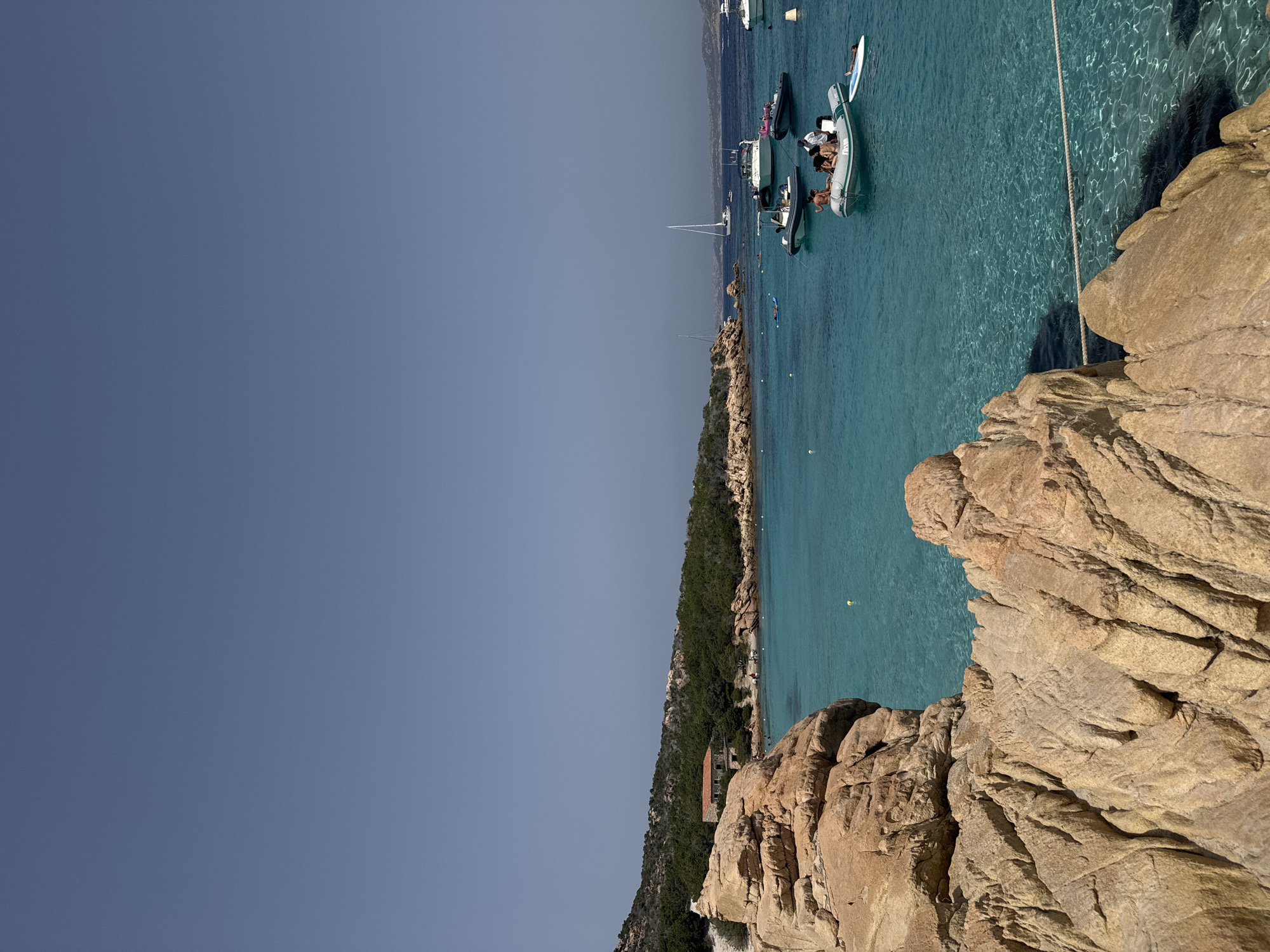 The image depicts a clear blue sky above a picturesque scene in Punta Sardegna, Italy. In the foreground, large, rugged, light-brown rocks dominate the left side of the frame, with textured surfaces and deep crevices. Beyond the rocks, a vibrant turquoise sea stretches out, its clarity allowing the seabed to be partially visible. Scattered across the water are several boats of various sizes, including inflatable dinghies with people in them and larger yachts. Some of the people are casually dressed, suggesting a relaxed summer atmosphere. In the middle ground, a small, red-roofed building is nestled among lush green trees and rocky terrain lining the shore. Further in the distance, other boats are visible, and the coastline curves into the background, dotted with vegetation. The overall impression is one of a sunny day in a beautiful coastal location, likely a popular spot for leisure and water activities.