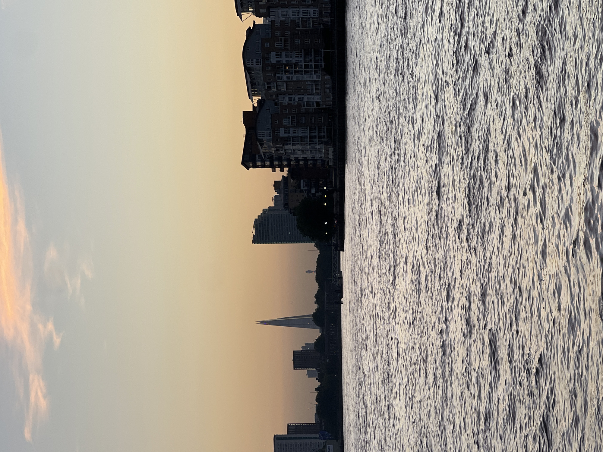 The image shows a skyline view of London from across the River Thames. The sky is a mix of orange and yellow, likely during sunset or sunrise. The Shard is visible in the skyline, along with other buildings of varying heights and designs. The river itself occupies a large portion of the image, with visible ripples and waves on the surface. The photo appears to be taken at dusk or dawn, with the buildings silhouetted against the sky.