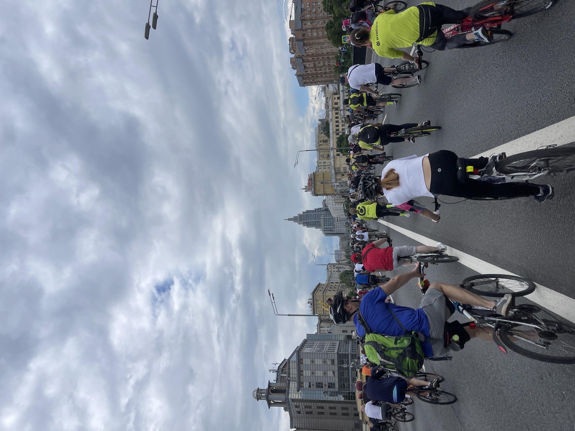 The image shows a large group of people riding bicycles on a road in Moscow, Russia. The cyclists are diverse in attire, with some wearing cycling gear and others in casual clothing. A prominent landmark, likely one of the Stalinist skyscrapers, is visible in the background. The sky is overcast, suggesting a cloudy day. The scene appears to be part of a cycling event or a group ride.
