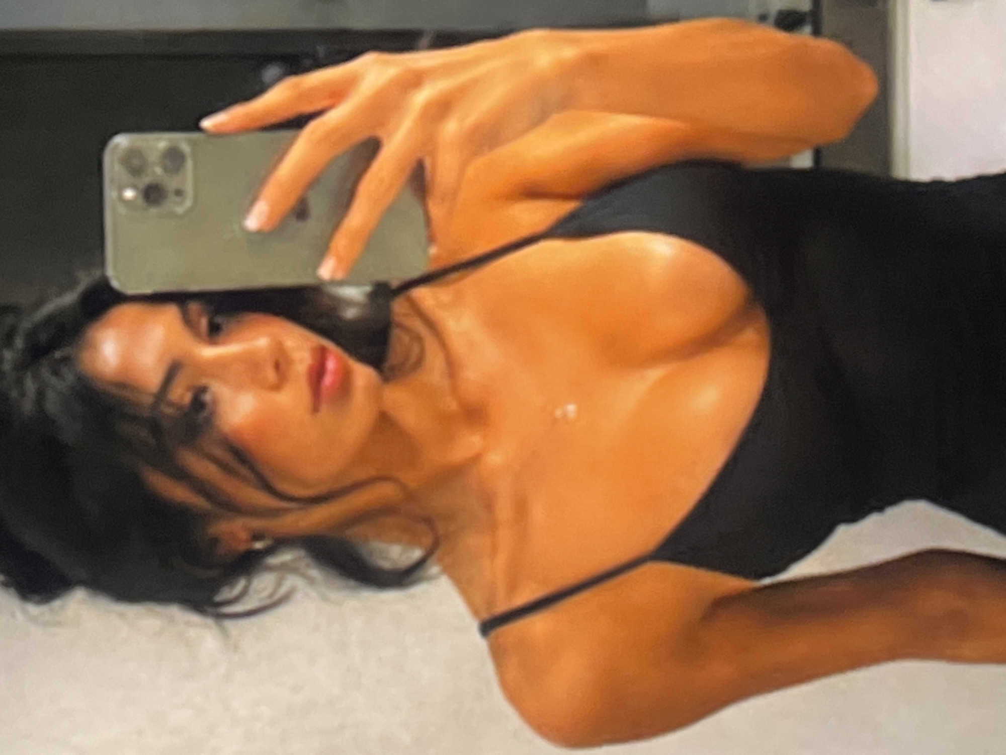 A young woman with dark hair styled in an updo is taking a mirror selfie. She is wearing a black spaghetti strap dress and a small silver necklace. Her right hand holds a light green smartphone, which is angled to capture the reflection. Her left arm is visible, bent at the elbow, with her hand resting on her hip. The background appears to be an indoor setting, possibly a bathroom, with a light-colored textured wall to her left and a darker, out-of-focus area behind her. The lighting suggests artificial illumination, casting a warm glow on her skin.