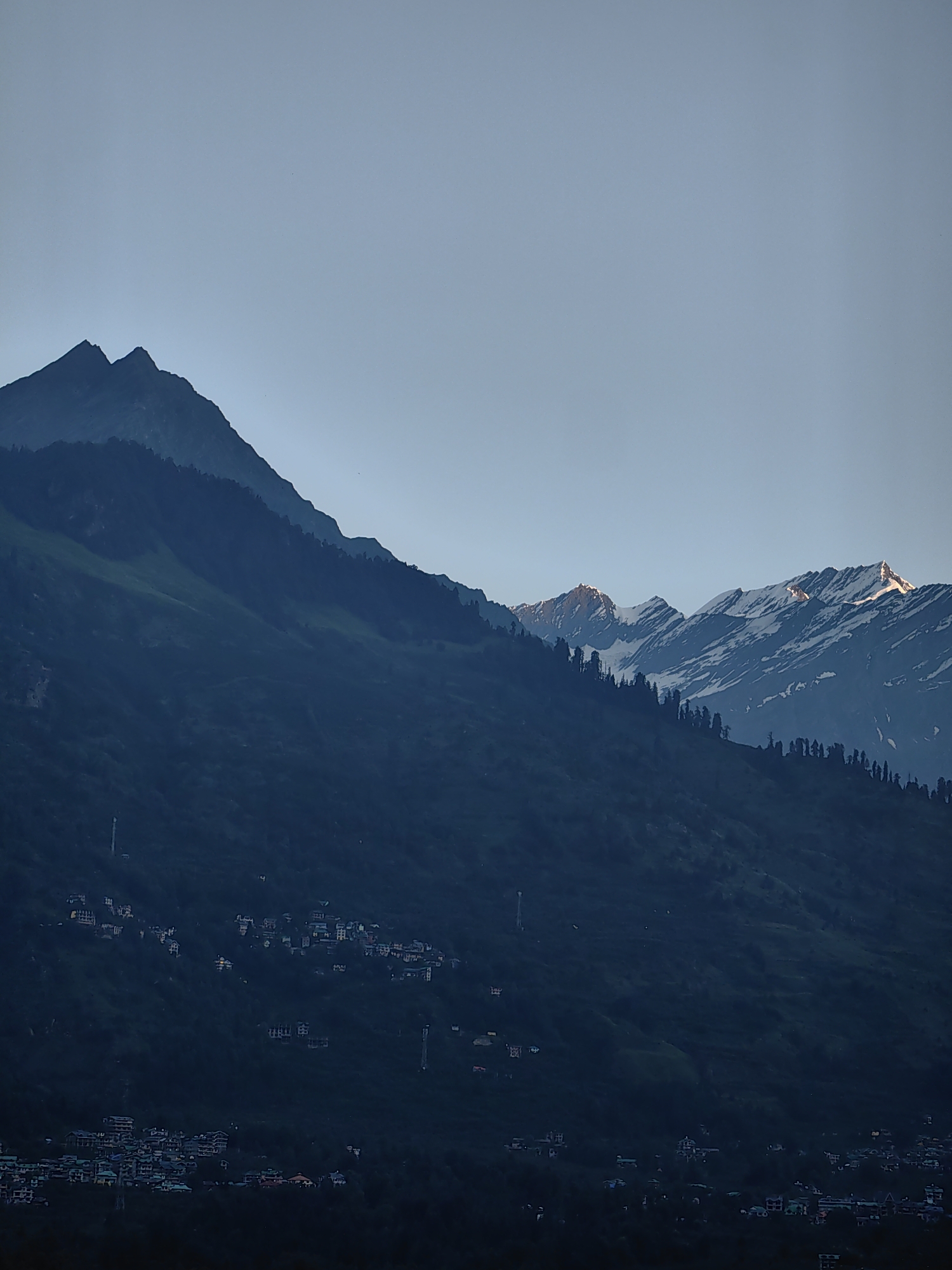 The image captures a vast mountain range under a clear, pale sky. The mountains are densely forested on their lower slopes, transitioning to rocky, snow-capped peaks at higher elevations. A small cluster of buildings, appearing to be a village, is nestled on one of the lower mountain slopes. The architecture of the buildings suggests a South Asian setting, with multi-story structures and pitched roofs. The overall impression is one of natural grandeur and human settlement in a mountainous region. The location is identified as Vashisht, India, a town renowned for its hot springs and mountainous surroundings.