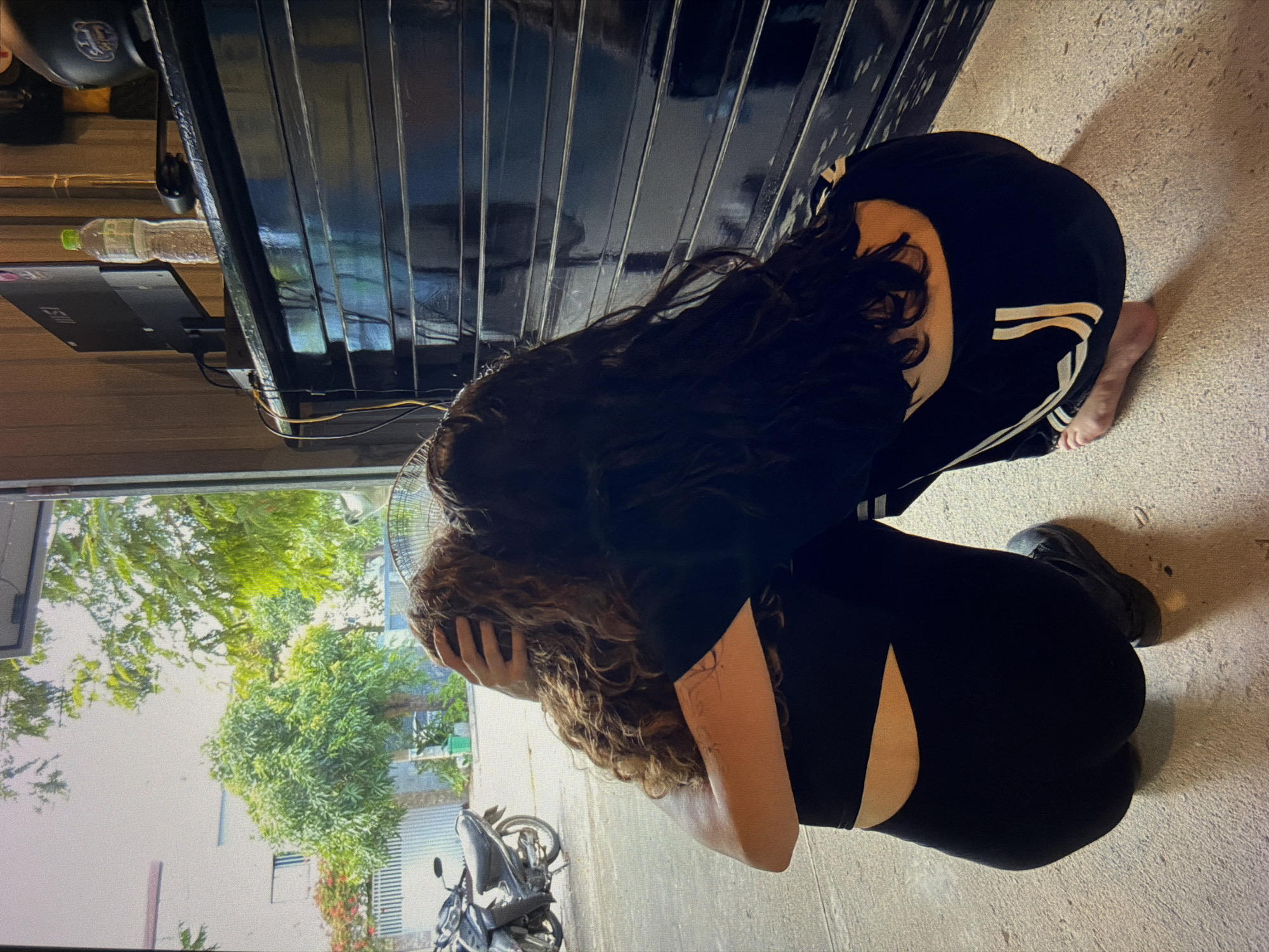 A photograph shows two people crouched on a concrete floor. One individual is wearing black leggings with a side cutout, a black shirt, and a single black shoe, with their head cradled in their hands. The other person is wearing black pants with three white stripes and a black shirt; this individual is barefoot. A motorcycle is visible outside, along with trees and buildings. Through the open garage door, an outdoor scene with trees and buildings is visible. A partially closed metallic garage door spans the background, with a bottle and electronic device sitting on the horizontal support beams. The scene is located in Đà Nẵng, Vietnam.