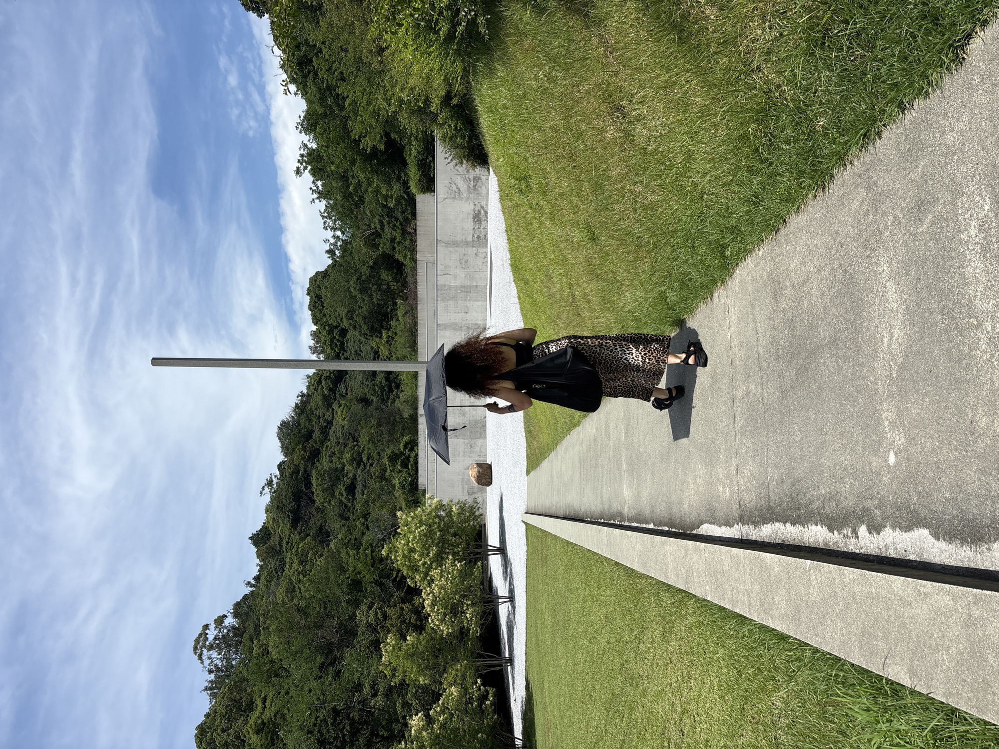 An individual walks along a paved path under a black umbrella. The person is facing away from the camera, wearing a leopard print skirt and dark sandals. They are carrying a black tote bag over their left shoulder. The path is bordered by a manicured lawn on the left and a grassy embankment on the right. In the background, a minimalist concrete structure with a tall, thin pole rises against a blue sky with wispy clouds. The area is surrounded by lush green trees and hills, suggesting an outdoor art installation or park setting. This is the outdoor space at the Chichu Art Museum on Naoshima Island, Japan.
