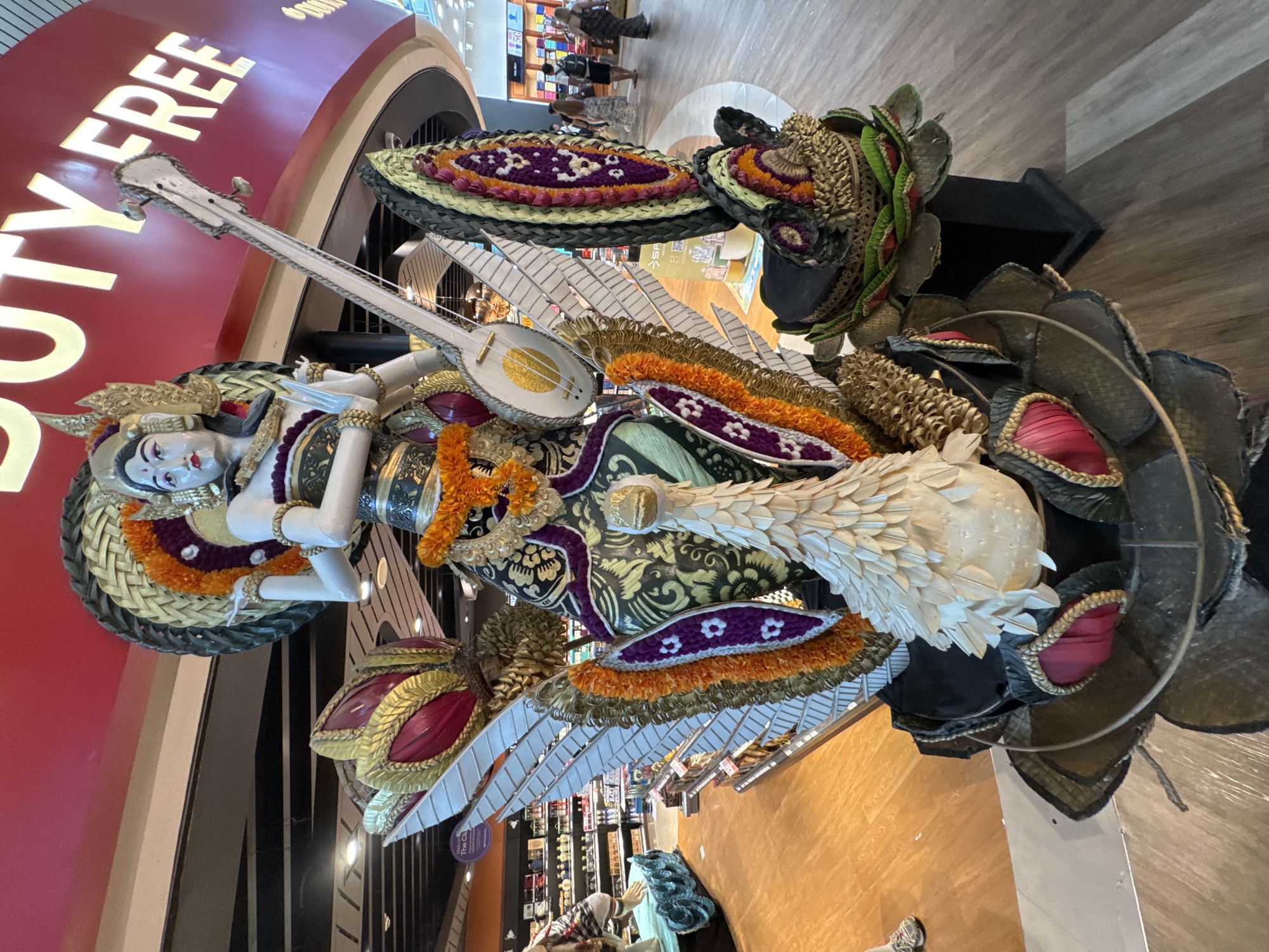 The image depicts a decorative sculpture in what appears to be a duty-free area within an airport or large retail space. The sculpture features a reclining female figure with pale skin and dark hair, adorned with ornate headgear and jewelry. She holds a white, lute-like instrument. The figure rests within a multi-layered structure of decorative elements, including flower arrangements, woven materials, and stylized representations of wings and petals. A sign with the words "DUTY FREE" in bold, capital letters is visible in the background. The setting suggests the sculpture is located in Kuta, Indonesia, due to the local style of the art piece.