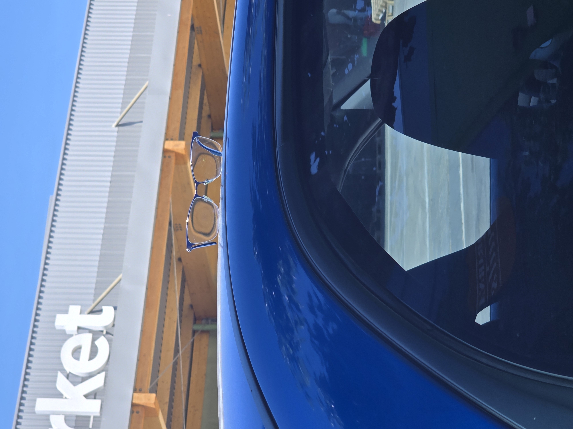A pair of blue-rimmed eyeglasses are perched on the edge of a blue vehicle, reflecting the facade of a building with the word "ket" visible, part of a larger word, likely "market." The building appears to be constructed with wooden supports and a corrugated metal exterior. The angle and reflection suggest the scene is outdoors on a sunny day. The location, Barcelona, Spain, is not evident from the visual details alone.