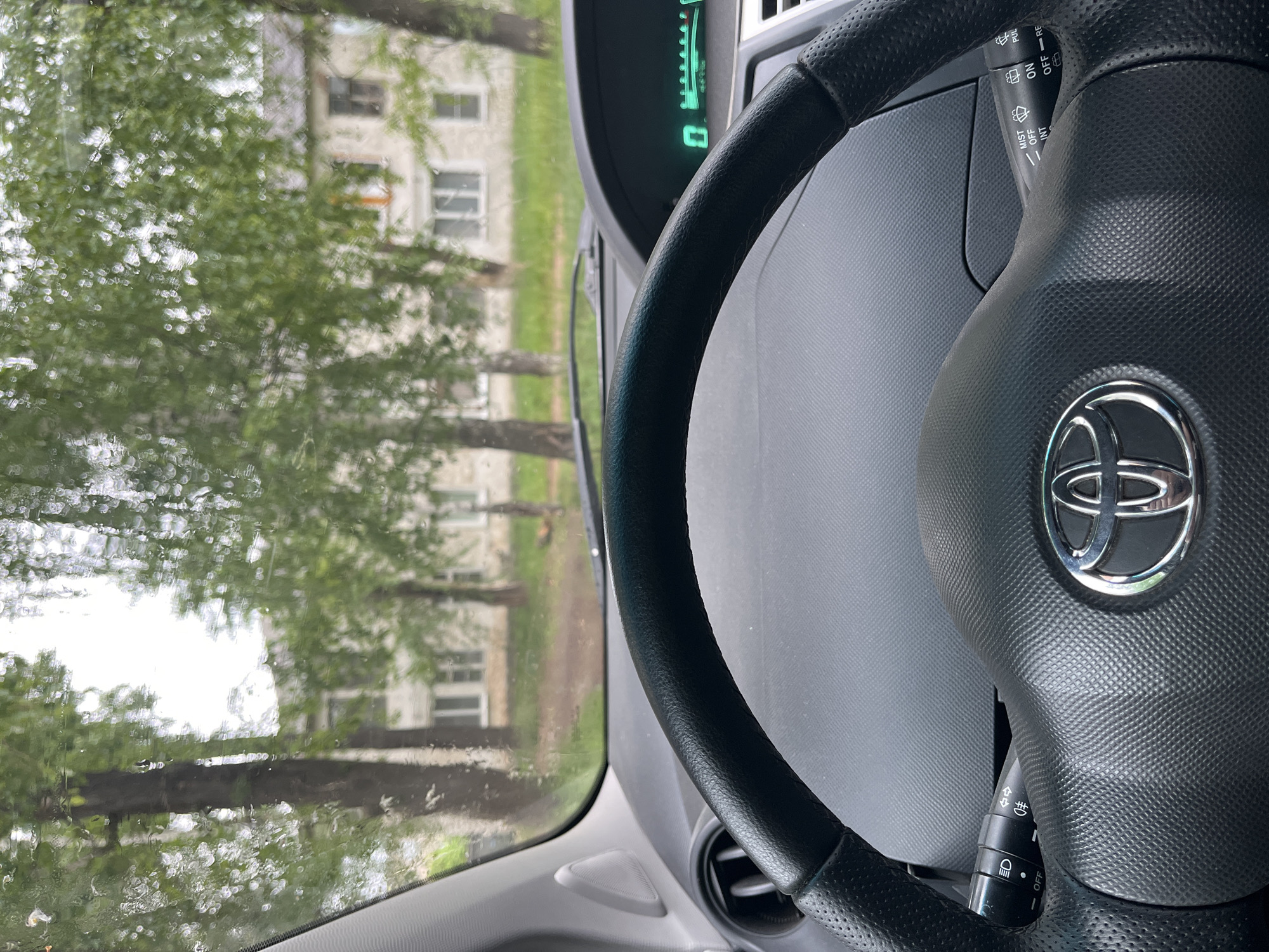 The image, taken from inside a vehicle, shows a perspective looking out through a rain-splattered windshield. In the foreground, occupying the right side of the frame, is a black, textured steering wheel with a prominent silver Toyota logo in its center. Behind the steering wheel, a digital dashboard display is partially visible, showing a green illuminated "0" (likely indicating speed) and several green bars, which could be a fuel gauge or battery level indicator.
To the right of the steering wheel, a stalk with controls for the windshield wipers is visible, featuring text such as "PULL", "OFF", "INT", "LO", "HI", "MIST", and "ON", "RE" (likely for rear wiper). Another stalk, presumably for lighting controls, is partially visible to the left of the steering wheel, showing symbols for headlights and fog lights, along with text like "OFF" and "DRL".
Through the rain-dotted windshield, an outdoor scene unfolds. Lush green trees with abundant foliage fill much of the view, creating a somewhat blurry, bokeh effect due to the raindrops and distance. Behind the trees, a light-colored, multi-story building with numerous rectangular windows can be seen. The weather appears to be rainy or recently rainy, as evidenced by the water droplets on the glass. The scene is illuminated by daylight.