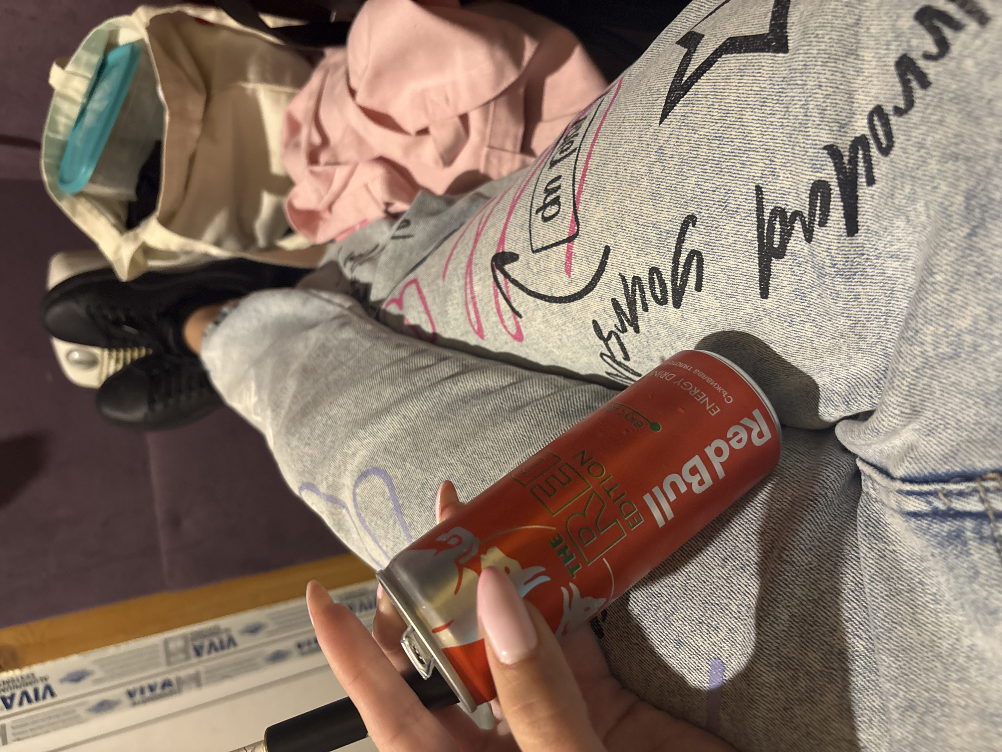 A person, likely female based on the fingernails, is holding a Red Bull energy drink can in their hand. The can is the Red Edition, which is cranberry flavored. The person is wearing light wash denim jeans with black writing on them. The jeans are on their legs, and the person is seated on a purple or dark upholstered surface. A white tote bag is visible near the top of the frame, with a pink garment on top of it. A pair of black shoes are partially visible near the tote bag. The scene appears to be indoors, with a window frame with "VIVA" markings on it. Without further information, it is impossible to confirm the location in Plovdiv, Bulgaria.