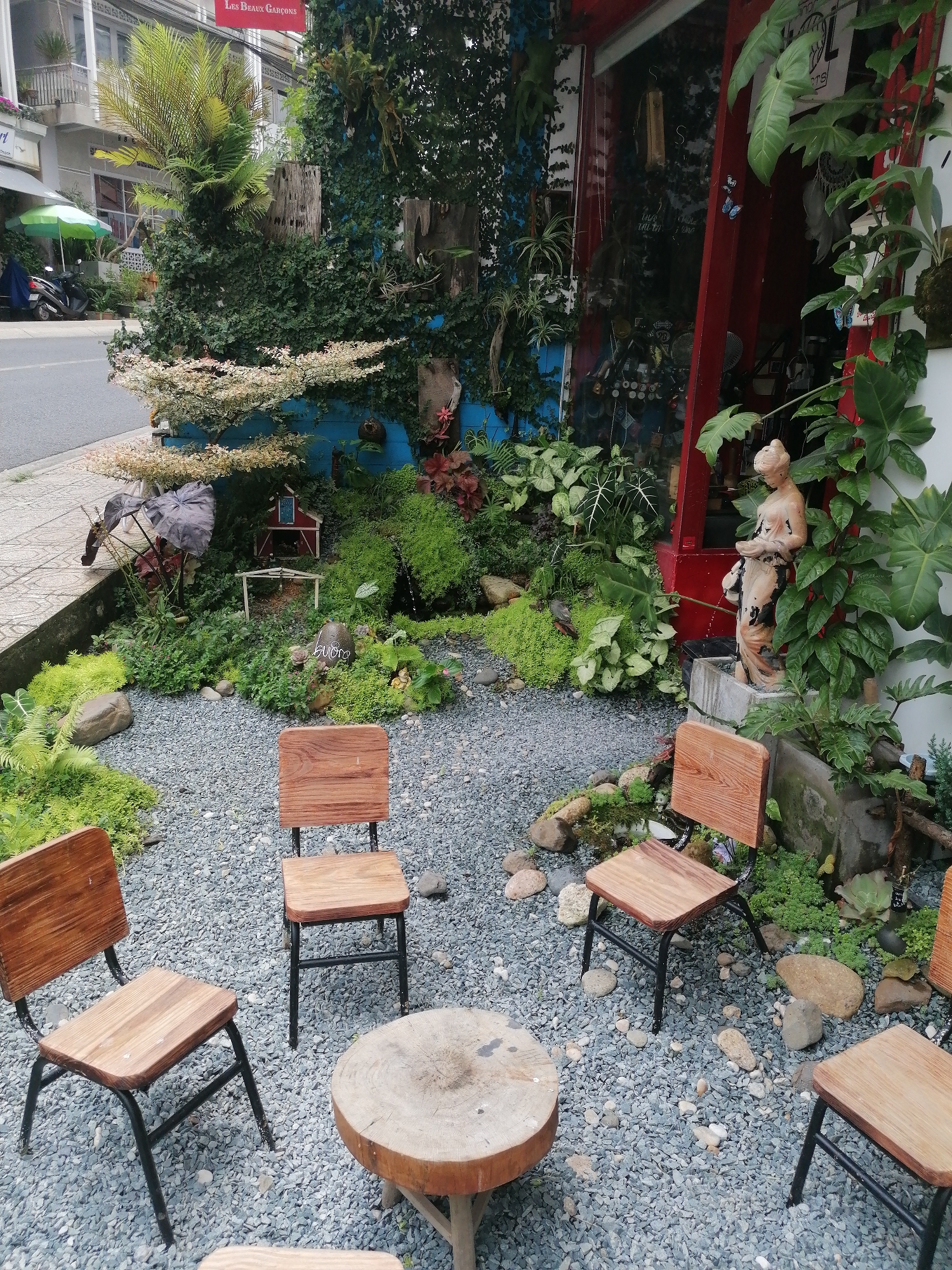This photo was taken in Da Lat, Vietnam. The image shows an outdoor seating area in front of a business. Three wooden chairs with black metal frames are arranged around a low, round wooden table on a ground covered in small grey pebbles. A fourth chair is partially visible on the right edge. The surrounding landscape is decorated with a variety of plants and shrubbery. A small statue of a woman stands beside the entrance, which has a red door frame. The building's exterior features a blue accent wall adorned with plants. A small ornamental house is nestled among the greenery. In the background, a street can be seen with a scooter parked under a green umbrella. A building with the sign "Les Beaux Garcons" is also visible in the background.