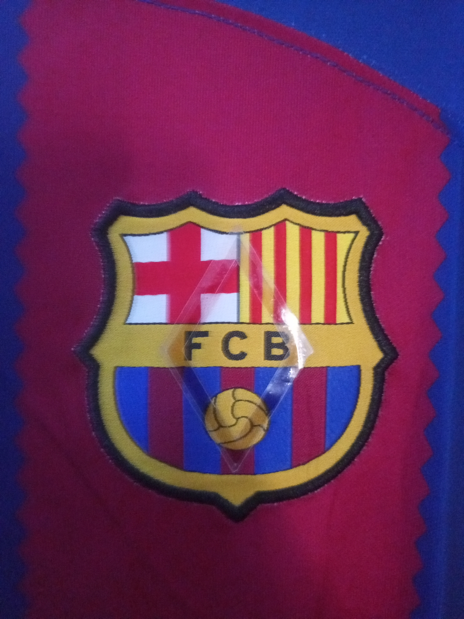 A close-up shot displays an embroidered FC Barcelona crest on a fabric, likely a sports jersey. The crest features the St George's Cross (red on white) and the Catalan Senyera (red and yellow stripes) at the top. Below these, the letters "FCB" are prominently visible. The bottom section of the crest shows vertical red and blue stripes, with a stylized, vintage-looking football centrally placed. The crest is outlined in black and yellow thread, giving it a raised, textured appearance.
A transparent, somewhat reflective, plastic or cellophane material covers the crest, creating a diagonal highlight across its surface. The fabric surrounding the crest is predominantly red in the central area, transitioning to blue on the left and right sides. The edges of the red fabric section exhibit a distinct zigzag or serrated pattern, suggesting an appliqué design or specific fabric cut. The overall texture of the fabric appears to be woven. No activity is depicted in the static image. While the location context is Maiburiji, Nigeria, there are no visual cues within the image to suggest this specific geographical setting.