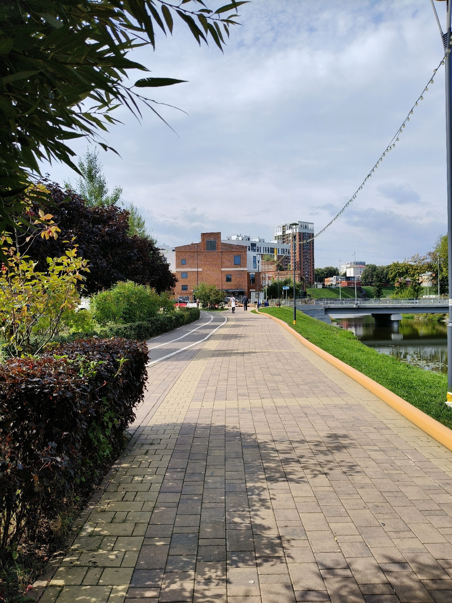 A paved pathway with a yellow border runs along a calm river, bordered by lush green grass and trees. On the left side of the path, there is a dense shrubbery with dark red leaves, and further back, a brick building with several windows and a modern multi-story building. On the right side, the river reflects the cloudy sky and the trees on the opposite bank. A bridge spans the river in the middle distance. Several people are walking on the path, and some are in the background near the buildings. The scene appears to be a public park or a waterfront promenade in an urban area, likely Naro-Fominsk, Russia, given the context.