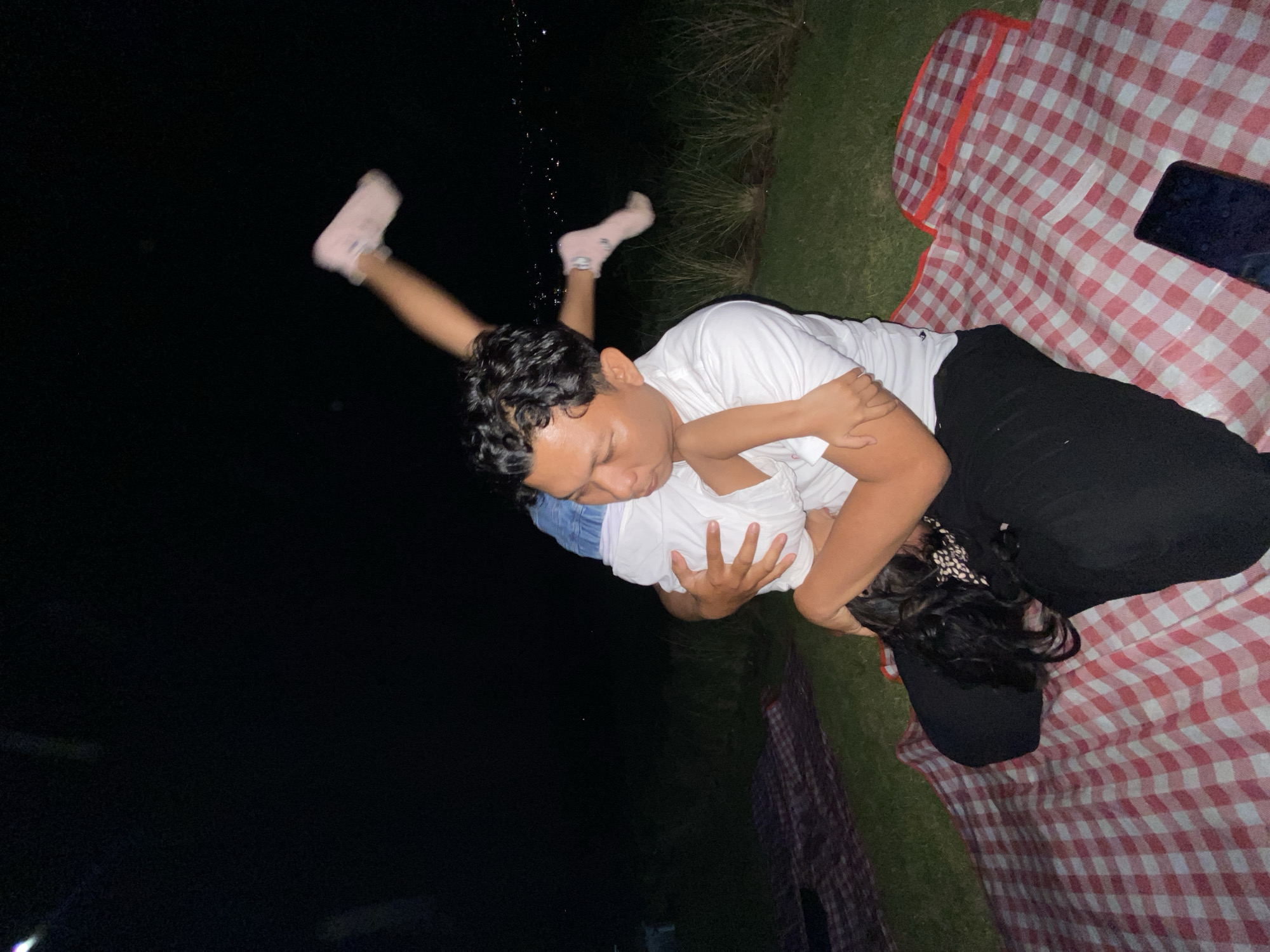 Two people are lying on a red and white checkered blanket, likely having a picnic in a grassy outdoor area at night. The person on top, wearing a white t-shirt and black pants, is holding the person underneath, who is mostly obscured but appears to have dark hair and is wearing a white shirt and possibly denim shorts. The person on top has their arm wrapped around the other person, and their faces are close together, suggesting an intimate or affectionate interaction. The person on top's legs are extended, with one foot in the air wearing a light pink sock. The background is dark, indicating nighttime, with some blurred greenery visible. The overall mood seems relaxed and possibly romantic. There is a phone on the blanket.