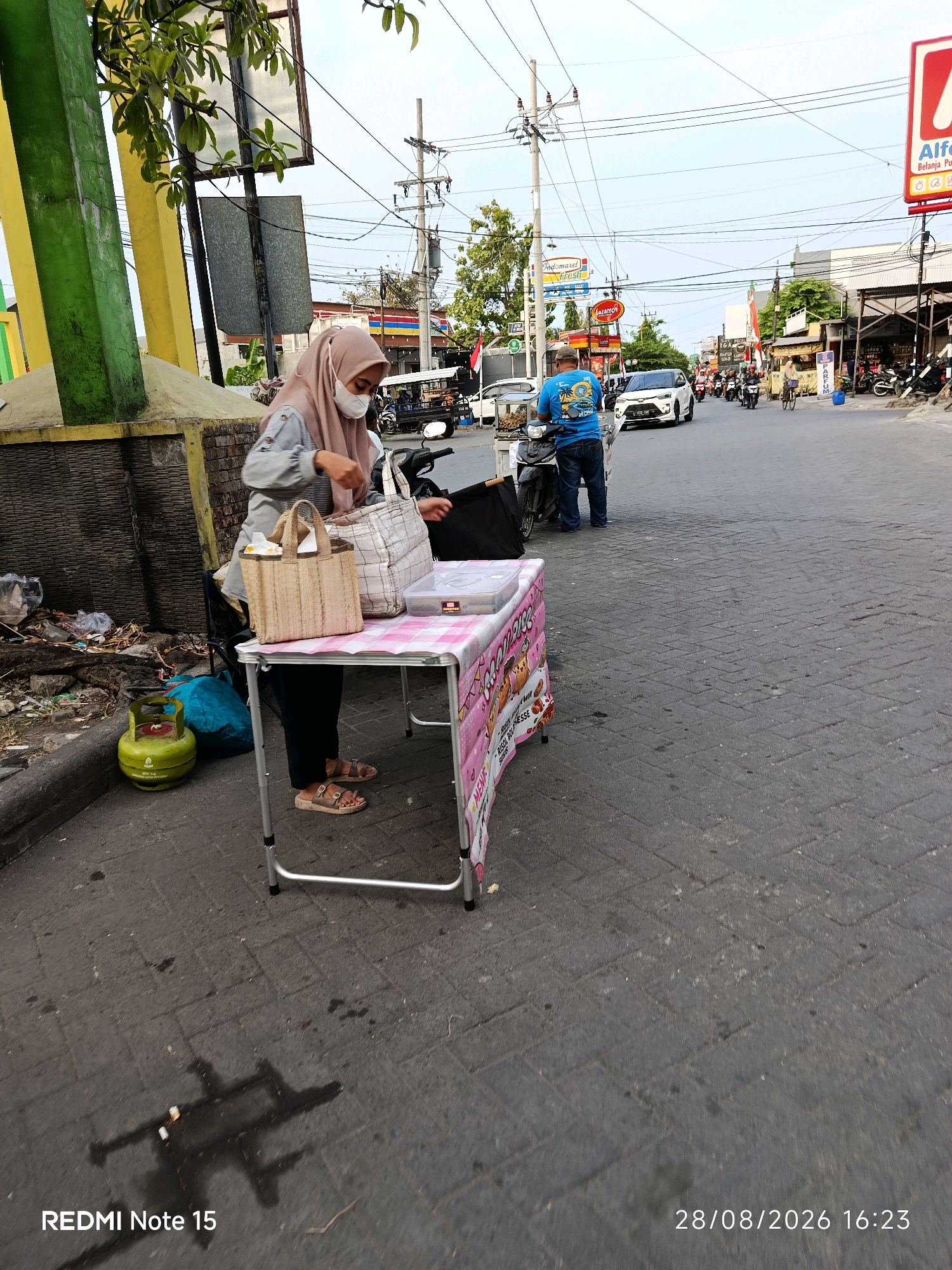 A medium shot captures a street scene in Keboansikep, Indonesia. In the foreground, a woman wearing a hijab and a face mask stands behind a small folding table covered with a pink and white checkered tablecloth. She is dressed in a light-colored long-sleeved shirt and dark pants, with tan sandals on her feet. On the table are two woven bags, one light brown and the other white and grey plaid, along with a clear plastic container. To the left of the table, a green gas cylinder is visible next to a pile of trash and a blue plastic bag. The ground is paved with interlocking bricks. In the middle ground, a man in a blue t-shirt and jeans stands next to a motorcycle. Several other motorcycles and a white car are on the street, with more vehicles further down the road. Buildings line the street, including storefronts with various signs. Overhead, power lines crisscross the sky, which is a pale blue. The overall impression is of a busy urban environment during daylight.