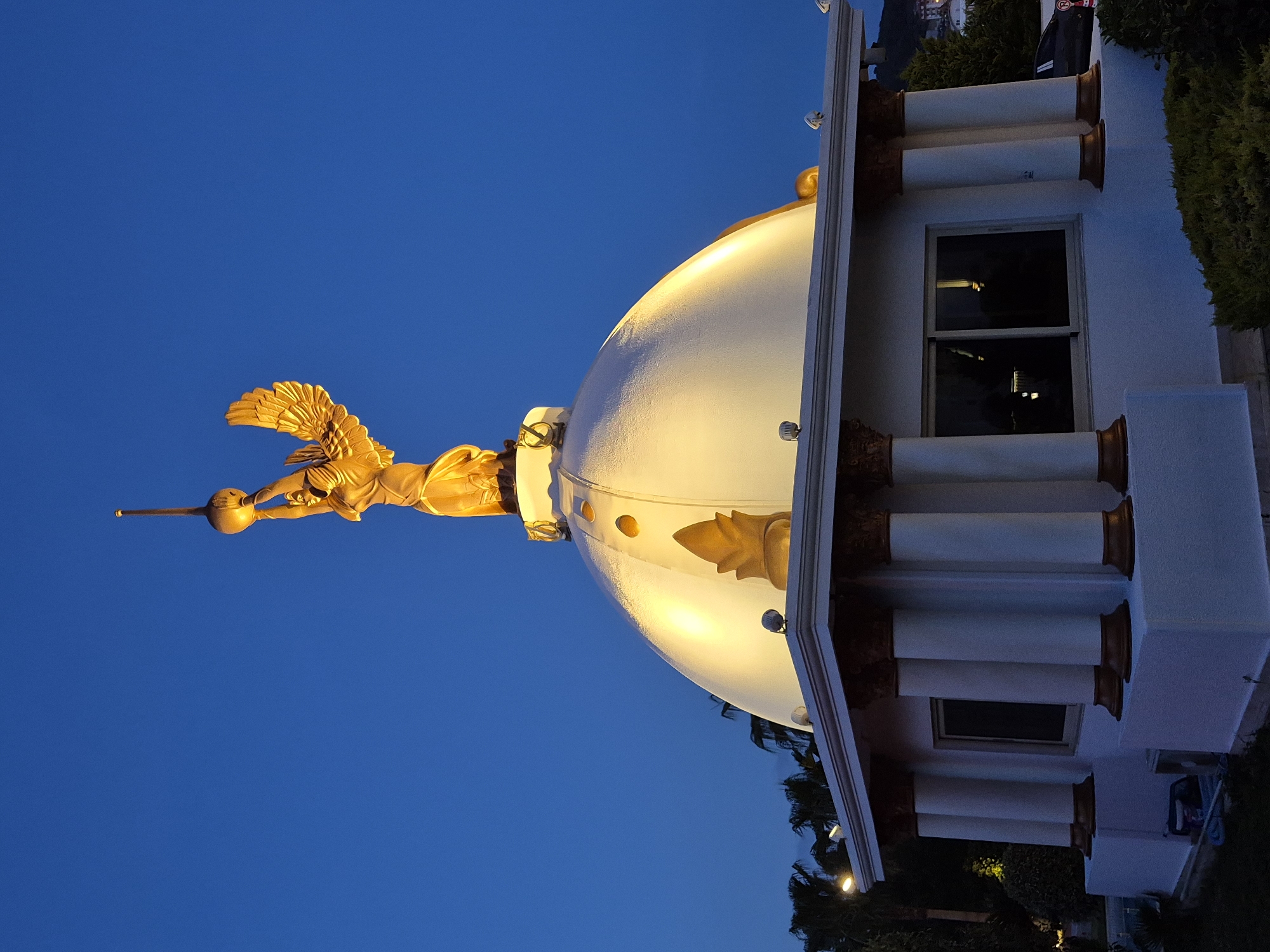 The image shows a low-angle view of a building topped with a golden angel statue against a blue sky. The building features a white dome and a white facade adorned with columns. The statue depicts an angel with outstretched wings holding a sphere on a long staff. The overall architectural style suggests a historical or cultural landmark. This building is the Temple of Love in Alanya, Turkey. There are bushes and trees flanking the right side of the building. A window is visible in the building's side facade.