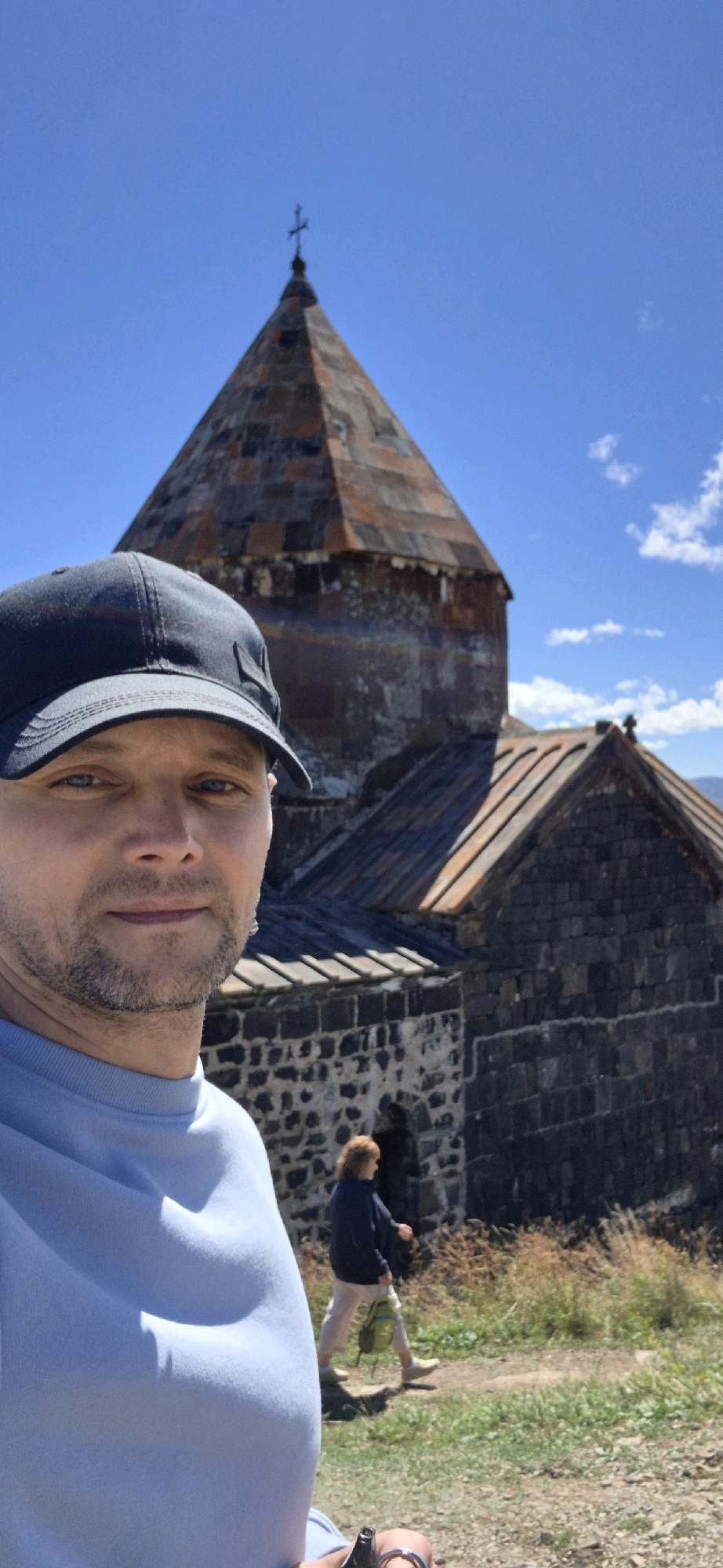 A man wearing a blue sweatshirt and a dark baseball cap takes a selfie in front of a stone church with a conical, weathered metal roof topped by a cross. The church appears to be made of dark, rough-hewn stones and is set on a grassy, slightly overgrown hill. The sky is a clear, vibrant blue with scattered white clouds. In the background, a person with reddish-brown hair walks away from the camera, wearing a dark jacket, light-colored pants, and carrying a green backpack. The scene suggests a visit to a historical or religious site, possibly on a sunny day. The church is likely part of the Sevanavank monastic complex located on the peninsula of Lake Sevan in the city of Sevan, Armenia, given its architectural style and prominent location.