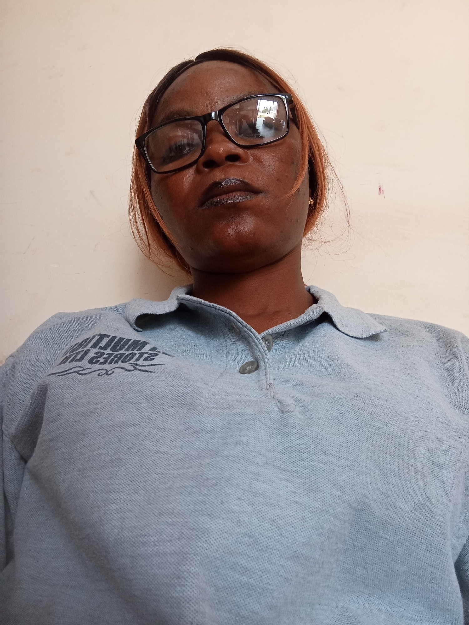 A woman with dark skin and shoulder-length reddish-brown hair wears black-rimmed glasses and dark lipstick. She is looking down at the camera, which is angled upwards, with her mouth slightly open. She is wearing a light blue collared polo shirt with a dark grey logo on the left side of the chest that reads "MJUM STORES". The background is a plain, light-colored wall. The image appears to have been taken indoors.