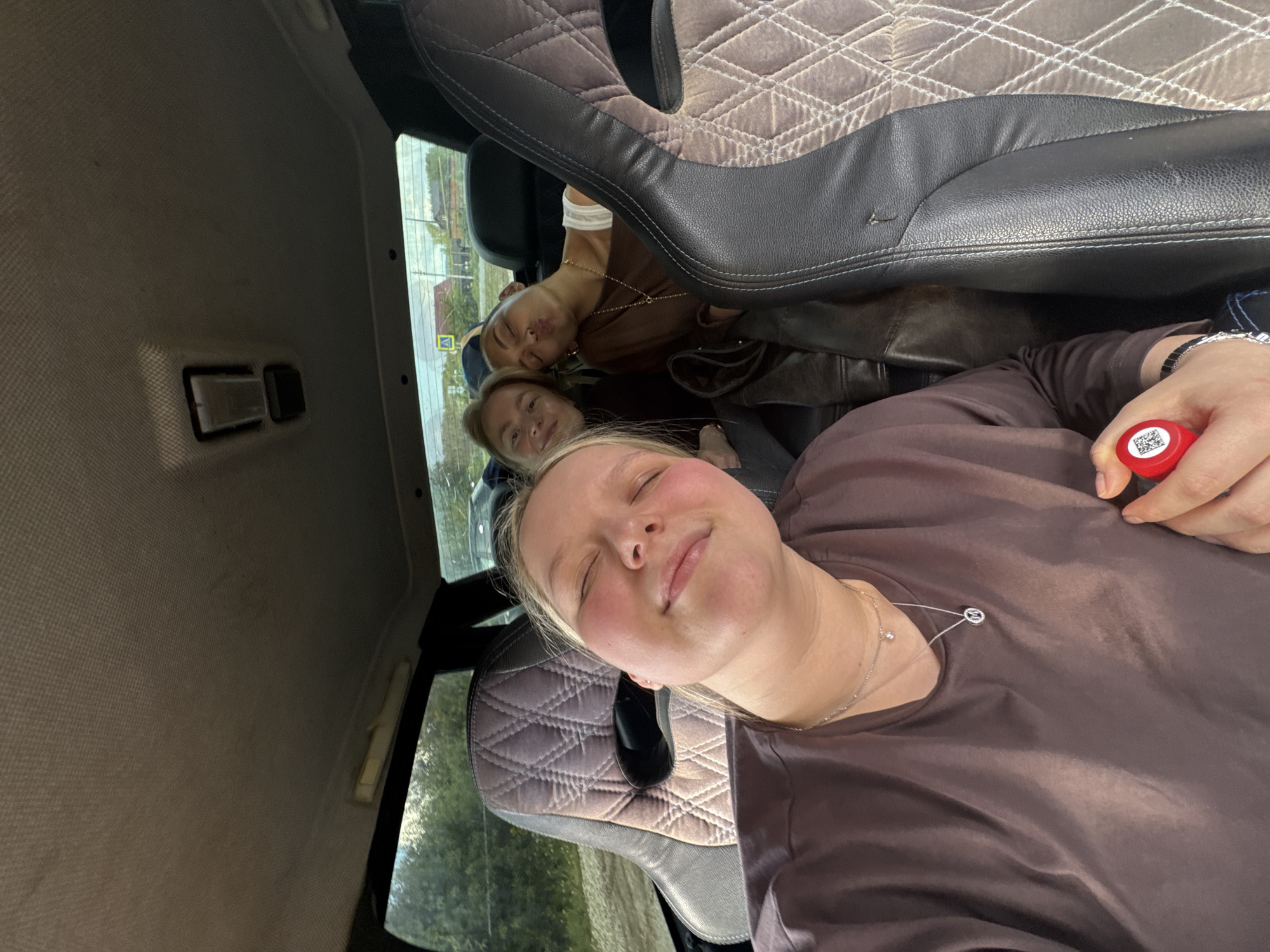Three light-skinned young women are inside a vehicle. The camera is positioned towards the woman in the foreground who is lying back on the passenger seat, facing upward with her eyes closed. She wears a brown satin top and a silver necklace. Her blonde hair is partly pulled back. A red bottle with a visible QR code is held in her right hand. Behind her, another woman with blonde hair looks toward the camera. In the back, a third woman is visible, appearing to rest her head. The car's interior includes patterned headrests and dark leather details. Through the car windows, a glimpse of a city is visible. A road sign, possibly indicating the location as Докша, Russia, is also visible.