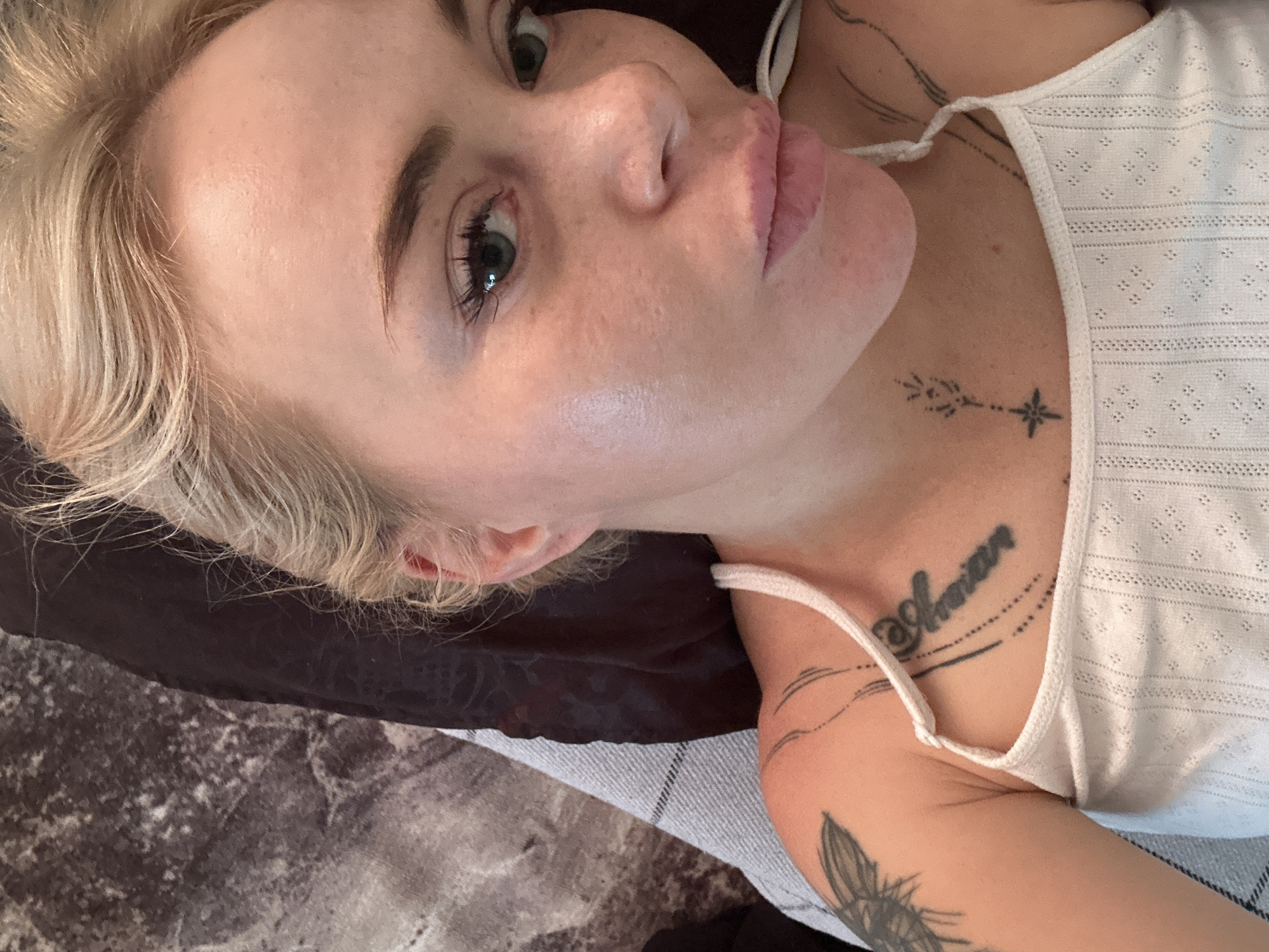 A close-up, overhead shot shows a young adult with blonde hair and light skin looking towards the camera. The subject is wearing a white tank top with a textured pattern. Tattoos are visible on the subject's left shoulder, upper chest, and neck. The background consists of a textured dark fabric and a grey and white striped surface. The overall setting is unclear, but appears to be indoors.