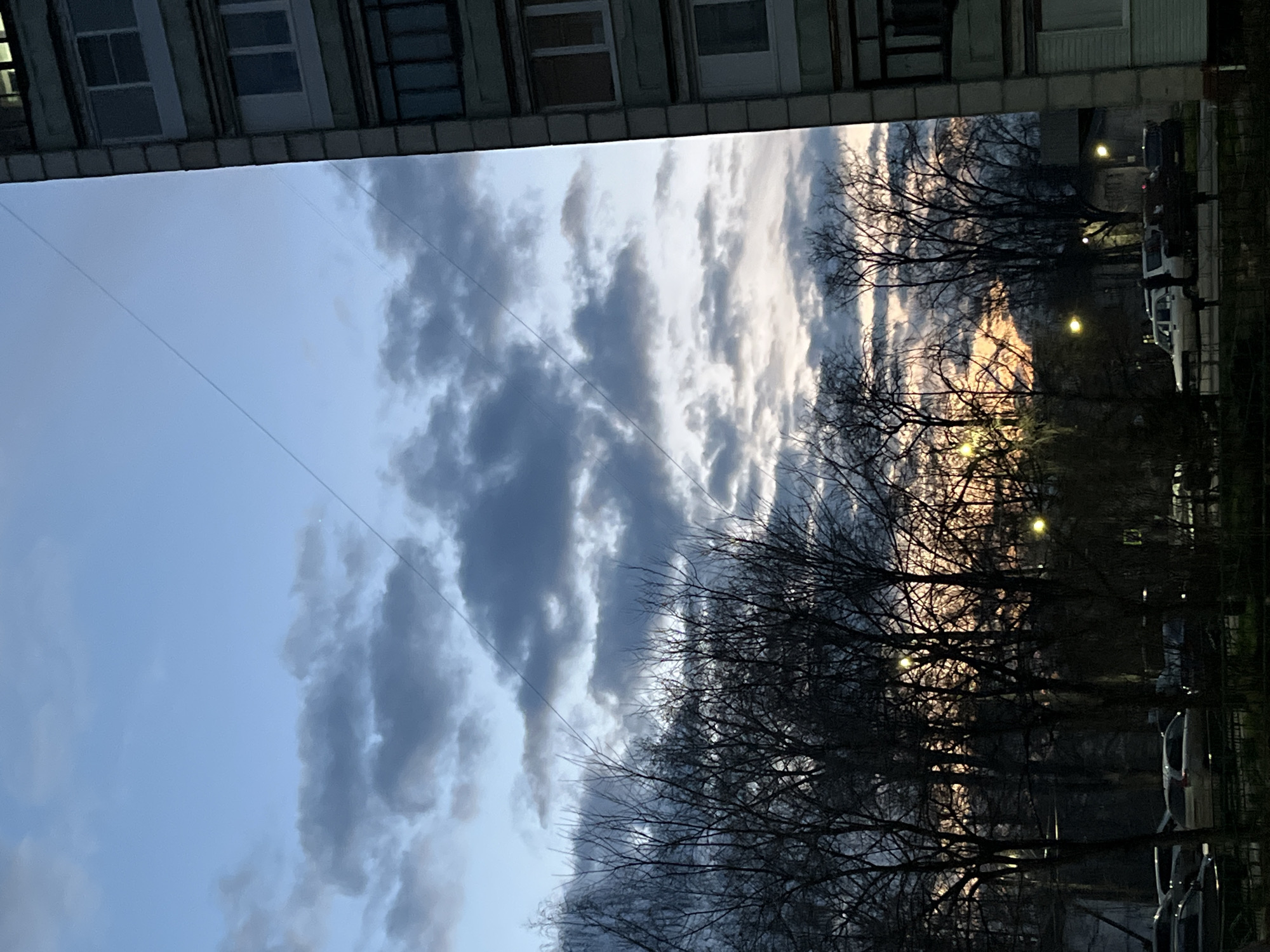 The image captures a view in Chaikovsky, Russia, looking upwards from what appears to be beneath the overhang of a building. The top of the image shows a section of the building, with several windows visible. The primary focus is the sky, which is filled with a mix of light blue and dark gray clouds. The overall scene seems to be taken during the early morning or late evening based on the sky’s coloration. Silhouetted trees with bare branches occupy the right side of the frame, adding a textured dark layer against the sky. In the background, beyond the trees, there are street lights illuminating what looks like a row of parked cars. Some power lines run across the sky in the upper left of the frame. There are no people visible in the frame.
