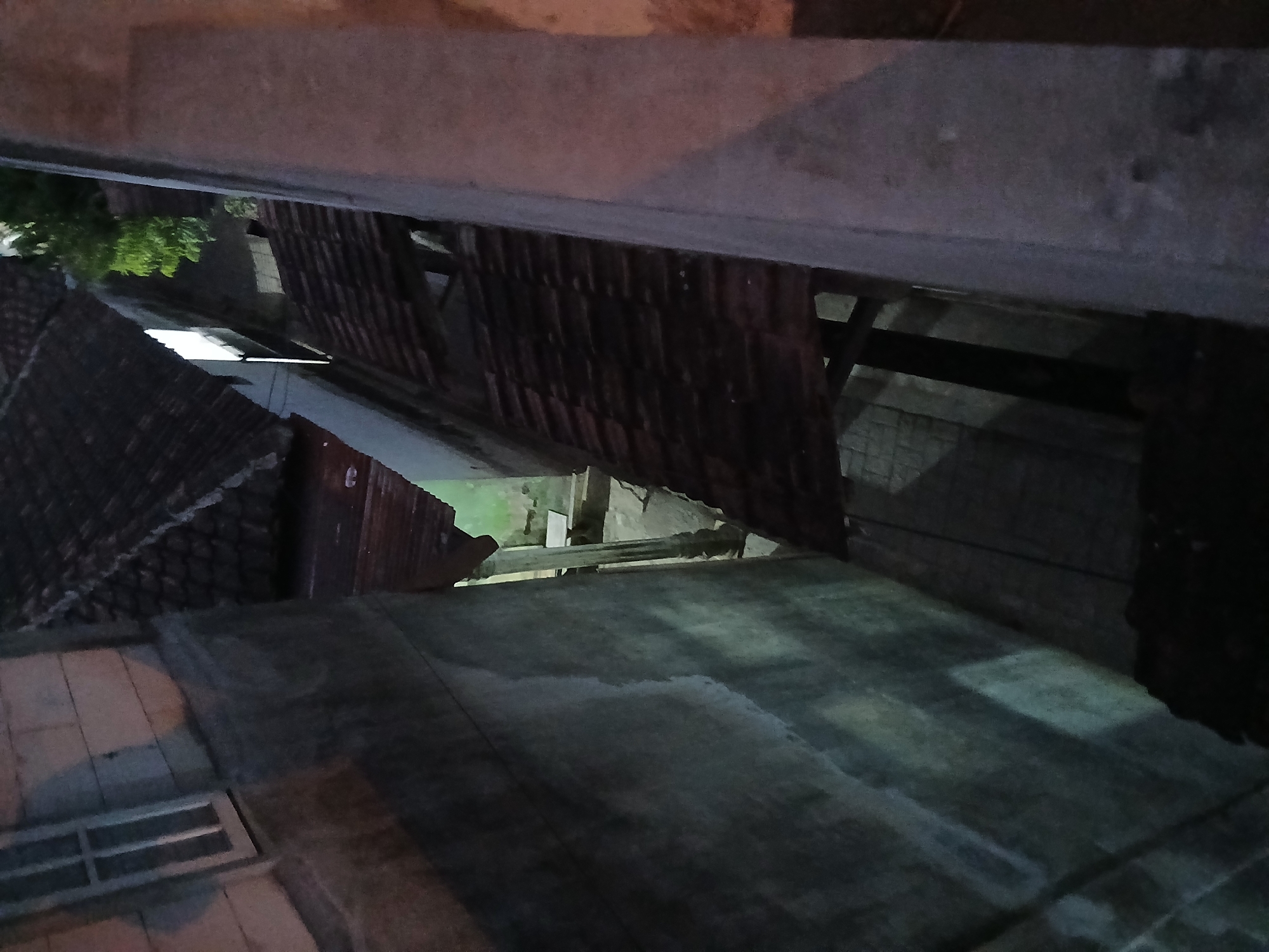 The image is a high-angle shot looking down a narrow alleyway between two buildings. The buildings have traditional tiled roofs and rough concrete walls. The alleyway is dimly lit, with shadows cast by the structures. There is a faint light emanating from an upper window of the building on the left. Lush green foliage is visible above the roofs in the distance, suggesting a residential area in Tenggarang, Indonesia. The scene appears to be a quiet, residential street at dusk or night.
