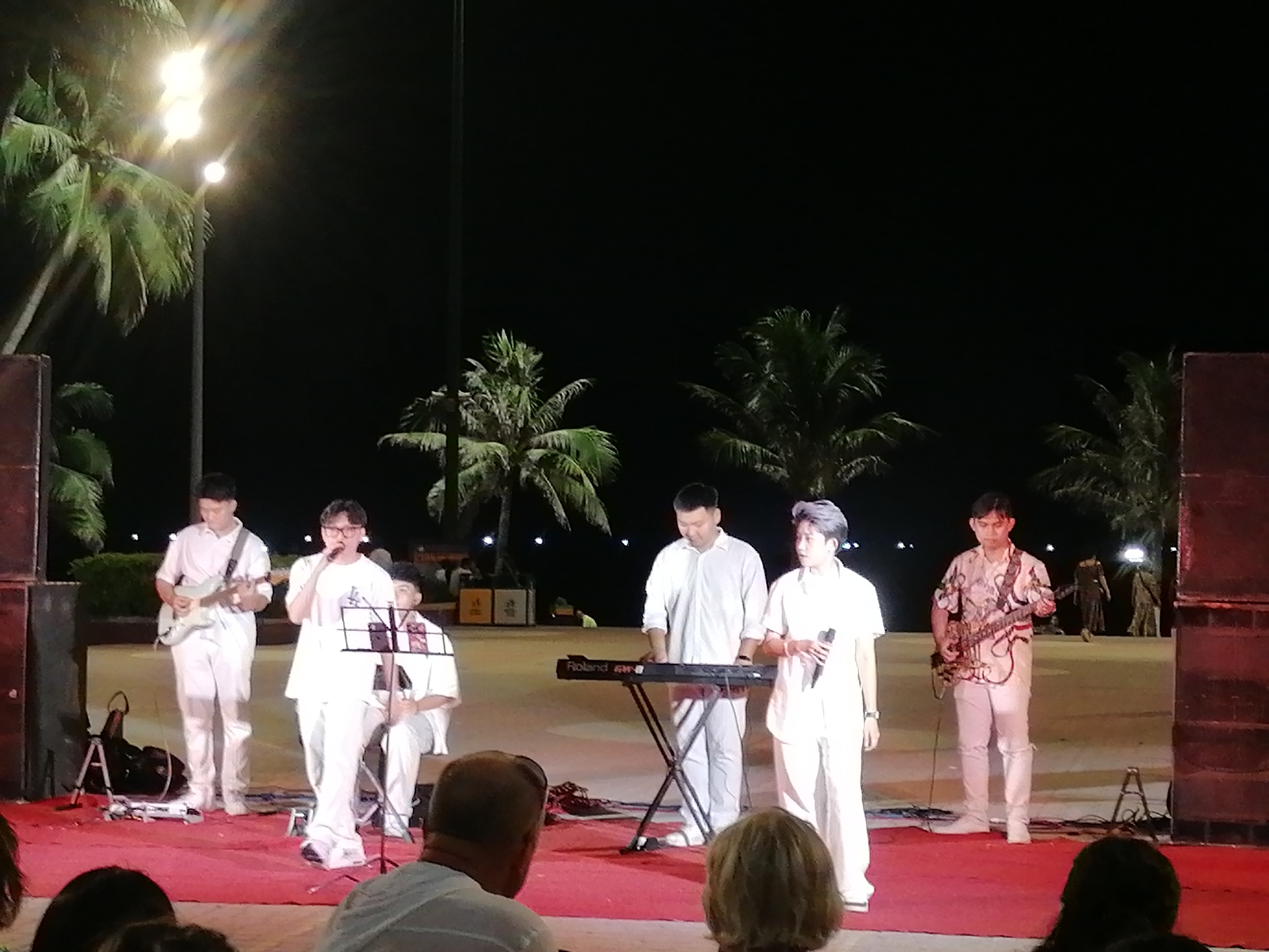 A band of five musicians performs on an outdoor stage at night, likely in the city of Nha Trang Ward, Vietnam. The musicians are dressed in white. One musician is playing an electric guitar, another is singing into a microphone with a music stand, another is seated, another is playing a Roland keyboard, and the last musician is playing a guitar. Palm trees can be seen in the background, along with stage lighting and what appear to be large speaker cabinets flanking the stage. An audience is seated in front of the stage.