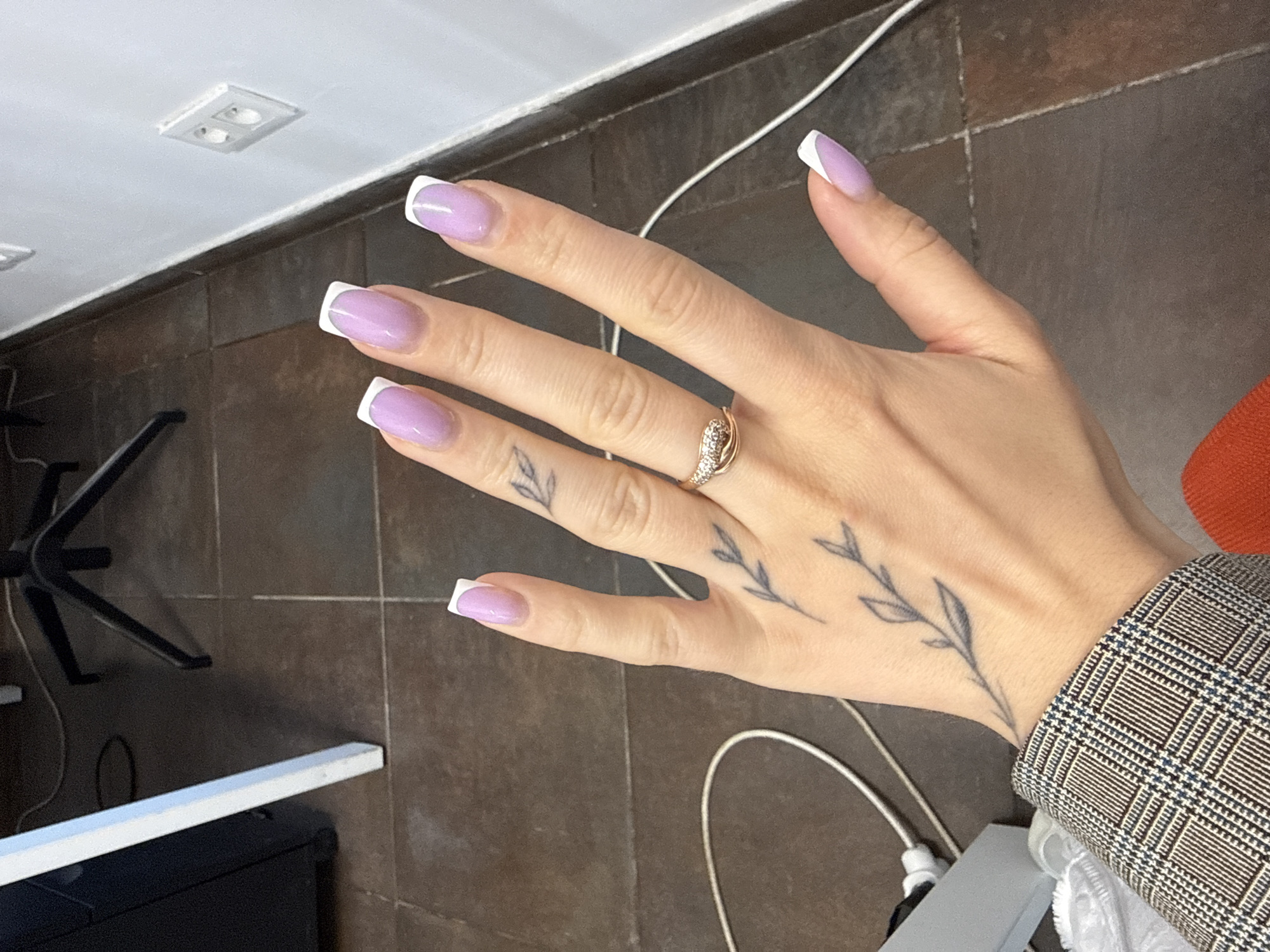 A person's hand with light purple French tip nails and a ring is shown against a brown tiled floor. The hand has a small black tattoo of a branch with leaves on the back. The background includes a white wall with electrical outlets, a black filing cabinet, and a chair base. A white cable is visible on the floor. The image appears to be taken indoors, possibly in an office setting, in Łódź, Poland.