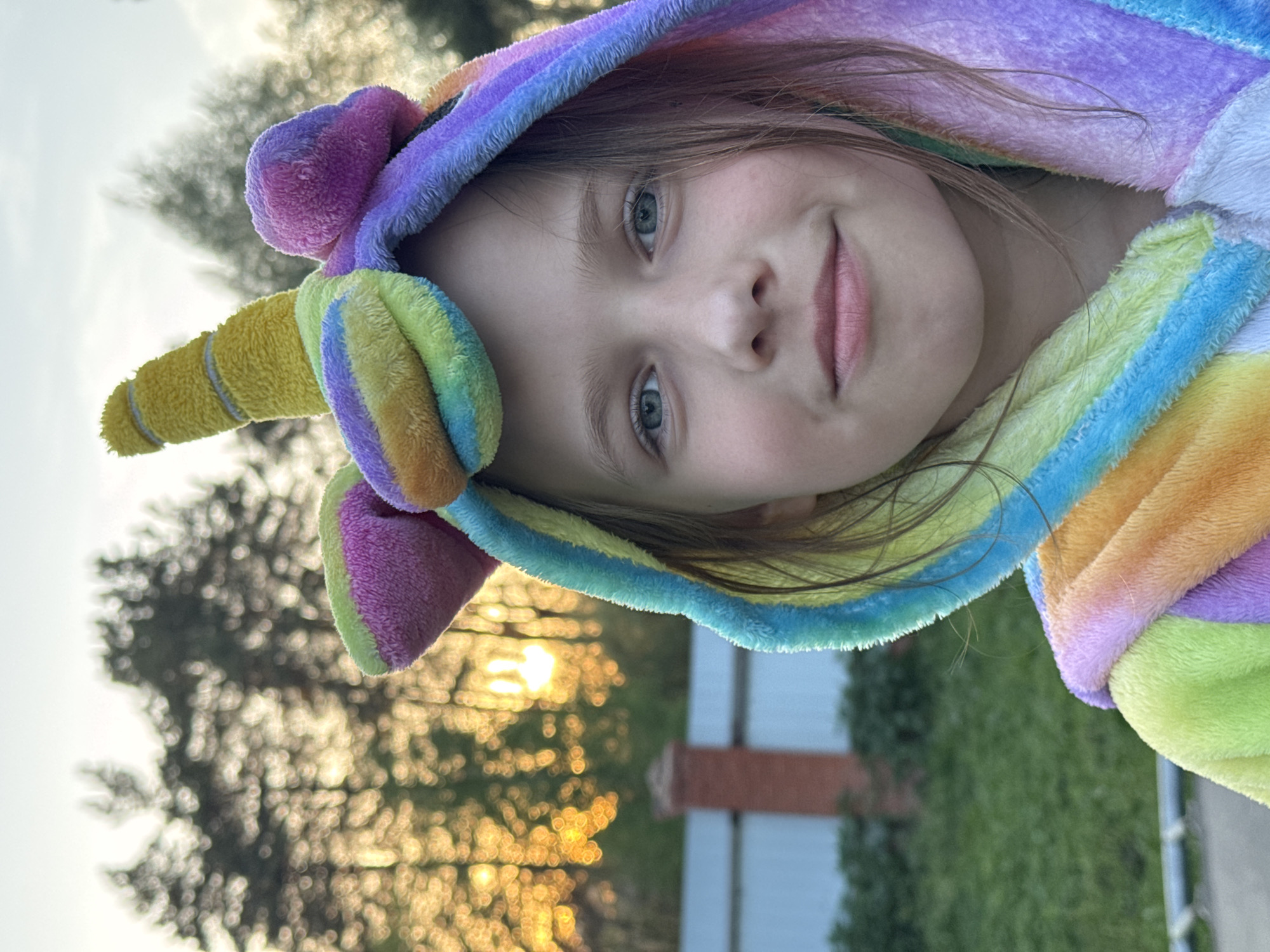 A young girl wearing a colorful unicorn hooded robe is the primary subject of the image. She is positioned in front of a blurred background consisting of trees, a fence, and a setting sun. The exact location within Rusalevka, Russia, cannot be determined from the visual information provided.