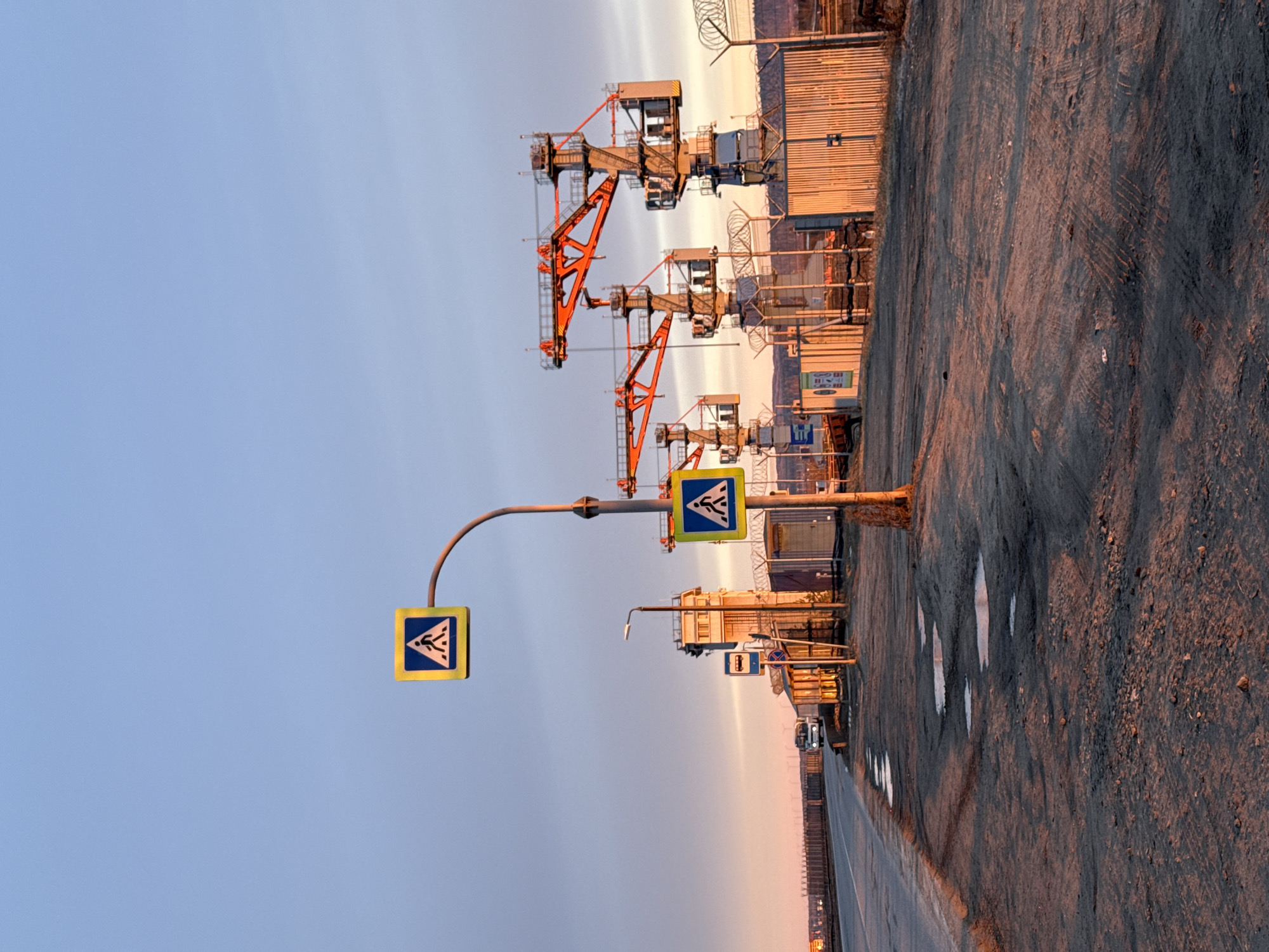 The image depicts a street scene in Chaikovsky, Russia at dusk. On the left is a pedestrian crossing sign attached to a curved pole, and another one further down the street. In the background, there are several metal structures, resembling cranes or towers, and containers. The ground appears to be dirt and gravel, with puddles reflecting the sky. The sky is a gradient of light blue to orange. There are no visible people in the image.