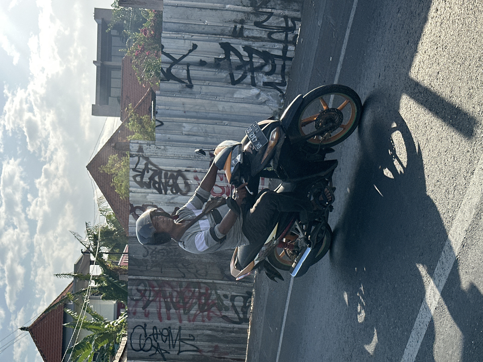 A man wearing a helmet rides a motorcycle on a paved road in North Kuta, Indonesia. The man is wearing a grey shirt with white stripes and dark pants. He has a dark bag slung across his shoulder. The motorcycle is black and orange, with a license plate visible that reads "DK 4342 DD". The background features a corrugated metal fence covered in graffiti. Behind the fence, buildings with red tile roofs and lush green vegetation are visible. The sky is partly cloudy, with sunlight casting long shadows on the road.