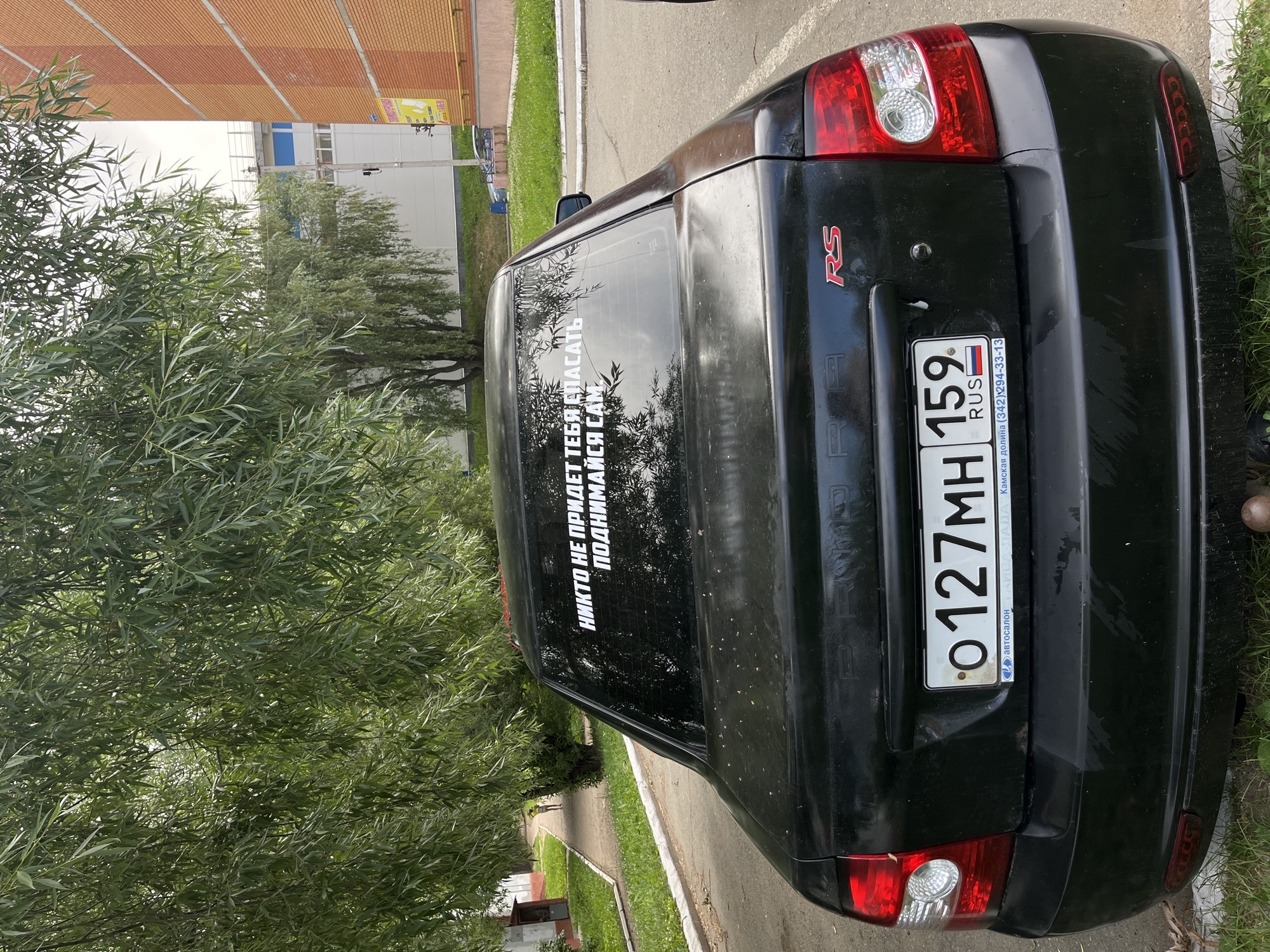 The image shows the rear of a black Lada Granta car parked on a paved surface in a residential area in the city of Chaikovsky, Russia. The car has a license plate that reads "O127MH 159 RUS". A white decal on the rear window says in Russian: "НИКТО НЕ ПРИДЕТ ТЕБЯ СПАСАТЬ ПОДНИМАЙСЯ САМ" which translates to "No one will come to save you, pick yourself up". The background features a sidewalk, grassy areas, trees with drooping branches, and buildings. The weather appears to be overcast.
