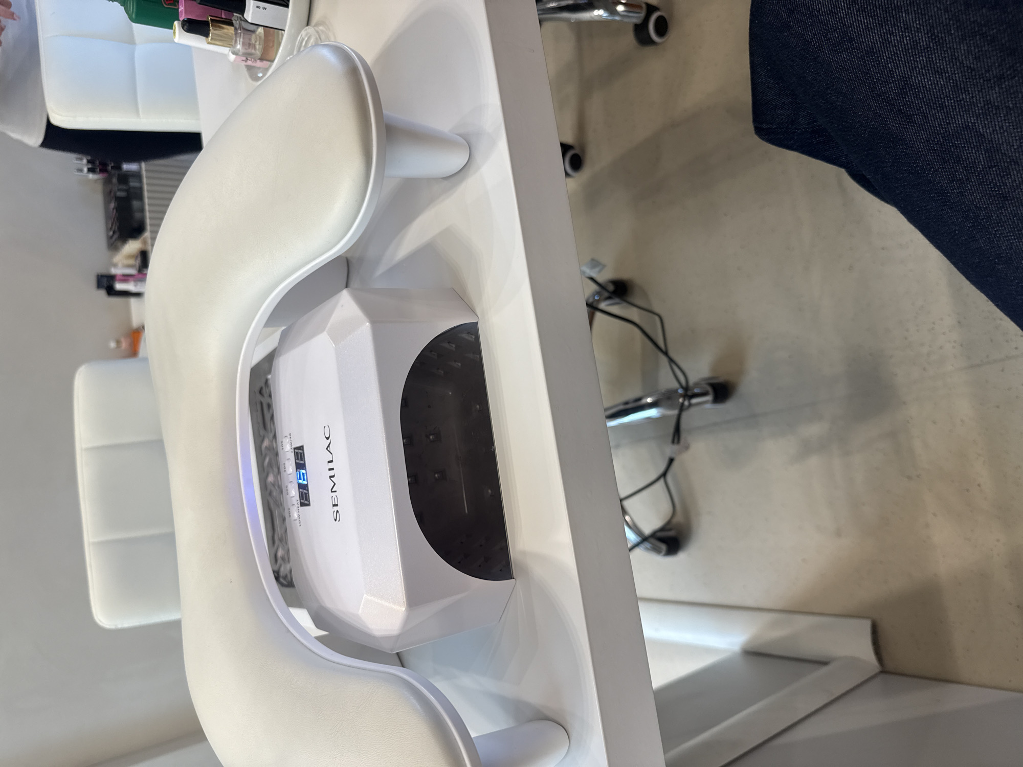 This image depicts a clean and modern nail treatment setup, likely in a salon in Milan, Italy, or a well-equipped home environment. The central subject is a white, polygonal UV/LED nail lamp, clearly branded "SEMILAC." Its digital display shows "888," presumably a timer, and it features multiple buttons for different curing durations: "5S," "30S," "60S," "99S," and a "LOW HEAT" option, with power specifications "24W/48" visible.
The nail lamp is placed on a sleek white workstation, which also features a custom-fitted, ergonomic white hand rest for client comfort. To the left, a portion of a white, upholstered chair is visible, indicating a seating area for a client. On the table in the background, several small bottles, likely nail polishes or other nail care products, are arranged, with "NO UV" partially visible on one label.
To the right, a person's leg, clad in dark blue denim jeans, is visible, suggesting someone is seated at the workstation, possibly undergoing or performing a manicure. Below the table, parts of a chrome-legged rolling chair with black wheels are visible, along with some electrical cables on the light-colored floor. The scene implies the activity of applying and curing gel nail polish. The lighting is bright and even, consistent with an indoor setting, and no specific time of day or weather conditions are discernible.
