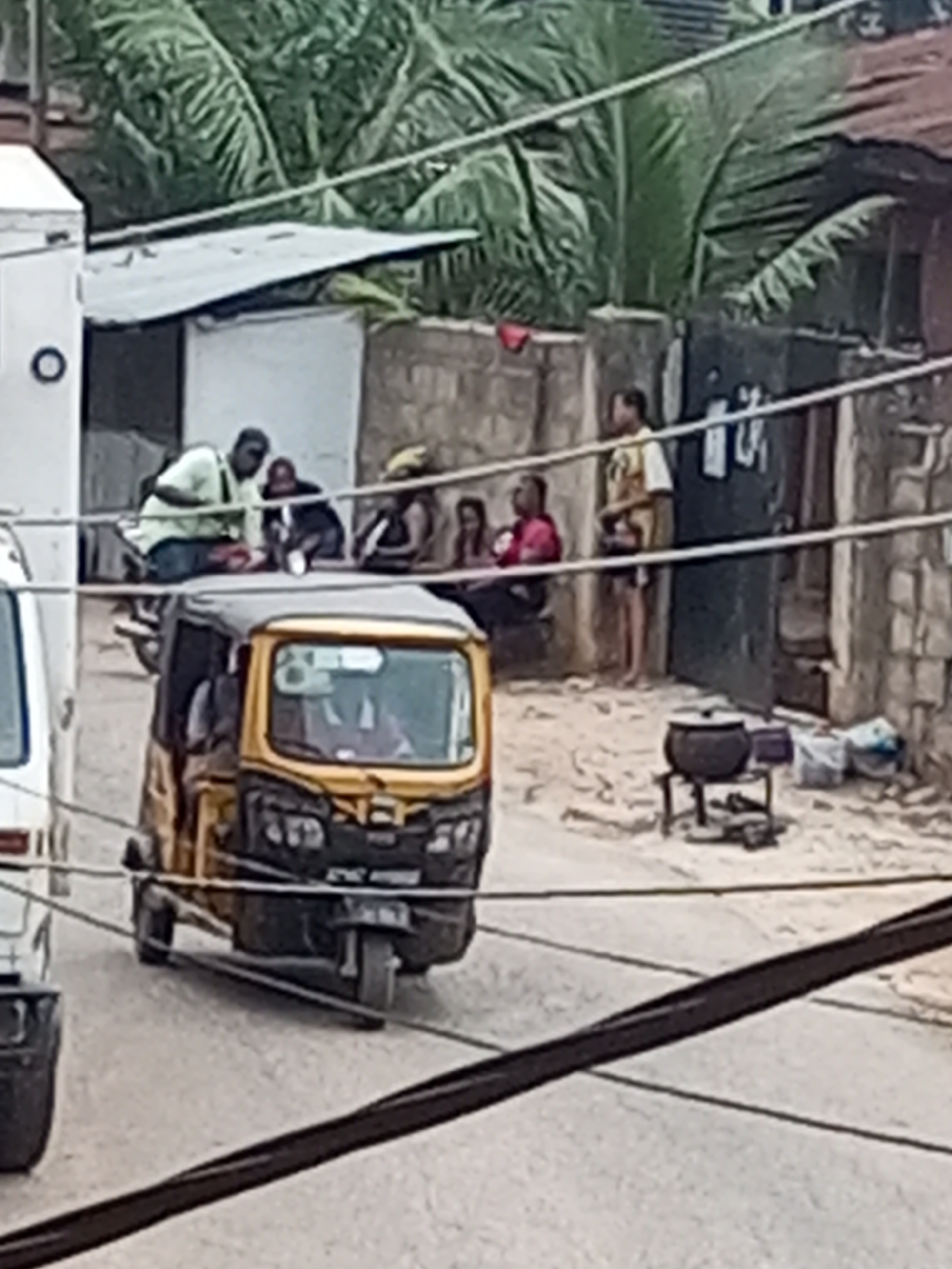 The image depicts a street scene in Onitsha, Nigeria. A yellow and black auto-rickshaw, known locally as a "keke," occupies the foreground of the image, partially obscuring a white van parked to its left. Several people are visible in the background, including a group seated or crouching against a concrete wall, and one man standing. A large, black cooking pot sits on a metal stand in the background. Utility wires crisscross the scene, partially obscuring the view. The environment includes palm trees and buildings with corrugated metal roofs.