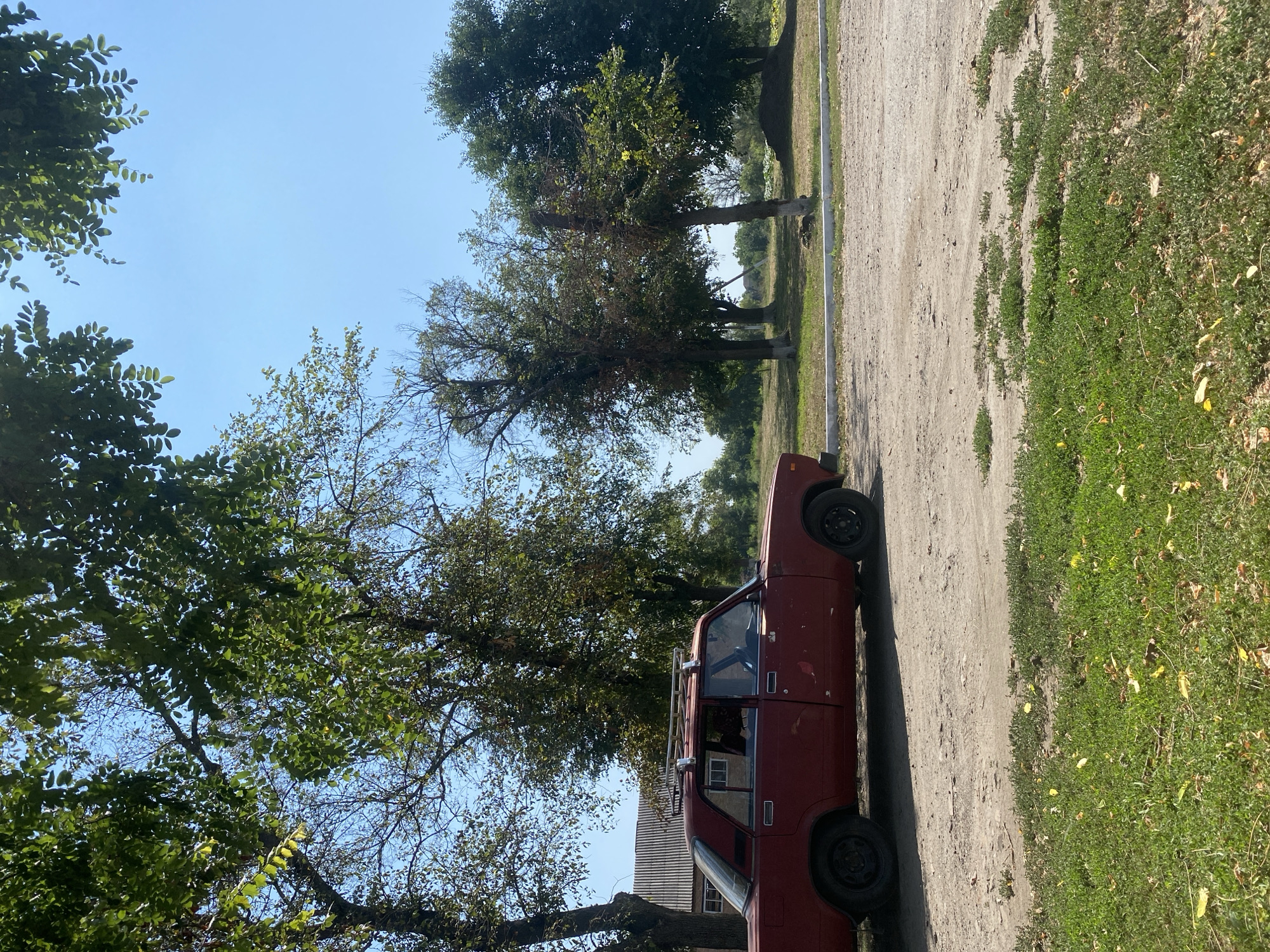 The image depicts a rural scene in the city of Ustyvytsia, Ukraine, on a clear, sunny day. A red, older model sedan, possibly a Lada, is parked on a dirt road. The car has a roof rack and its windows are tinted or the interior is dark. A person can be seen through the driver's side window. To the left of the car is a building with a corrugated metal roof, suggesting a utilitarian structure like a barn or shed. The surrounding environment is characterized by several trees, some with lush green foliage and others with sparser, drier branches. The ground in the foreground is a mix of dry dirt and grass, with scattered yellow leaves. In the background, more trees and vegetation are visible, with a clear blue sky above. The overall impression is of a quiet, perhaps slightly neglected, rural location.