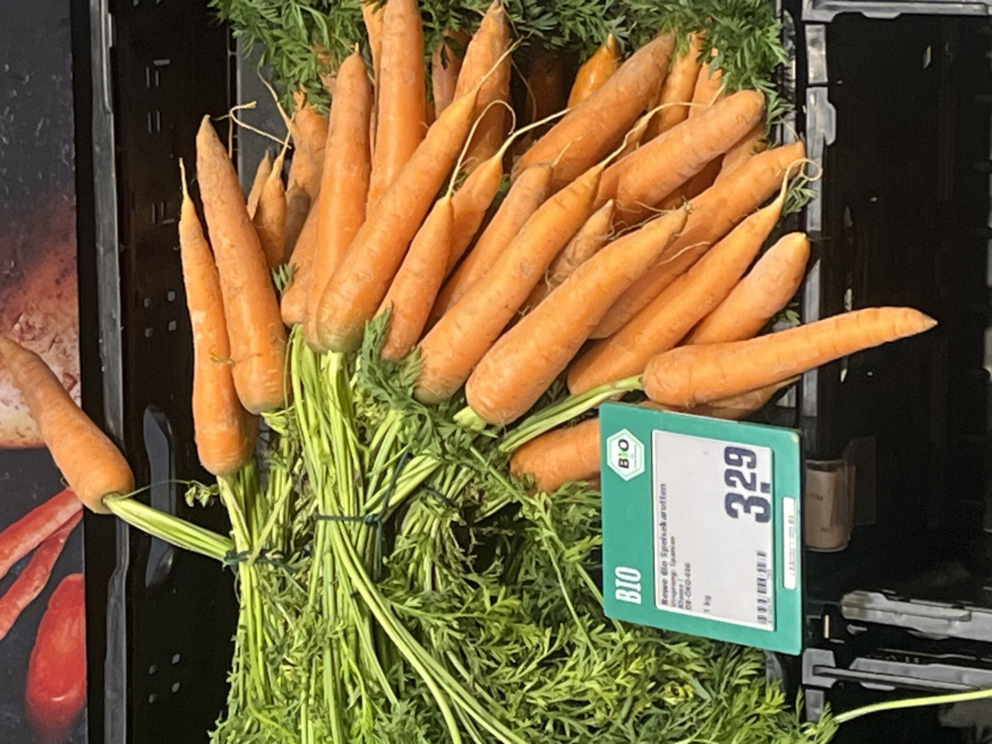 A bunch of fresh, orange carrots with green tops is displayed in a black plastic bin. A price tag is attached to the bin, showing "Rewe Bio Speisekarotten" and the price "3.29". The carrots appear to be organic. The background suggests a grocery store setting, possibly a Rewe supermarket in Berlin, Germany, given the presence of the Rewe brand. The lighting is bright, indicating an indoor commercial environment. There are no people or animals visible in the image.