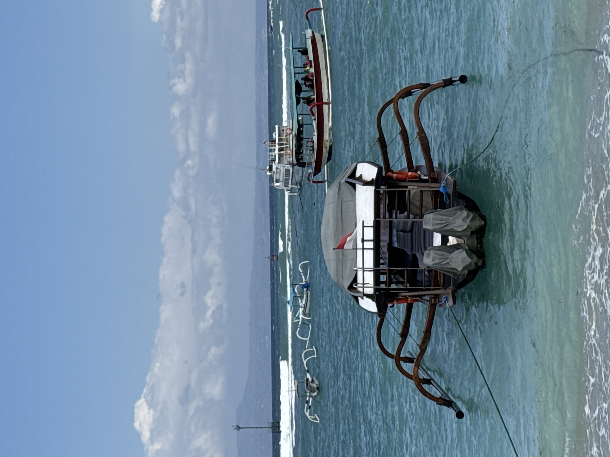 The image depicts a fleet of boats anchored in the turquoise waters off the coast of Manggis, Indonesia. In the foreground, a traditional Indonesian fishing boat, known as a "jukung," with its characteristic outriggers, is tied to the shore. The boat is partially covered by a grey tarp, and two large outboard motors are visible at the stern. A small Indonesian flag flies from a pole on the boat.
Further out in the water, several other jukungs are visible, some with passengers. In the middle ground, a larger white ferry is also present. The sea is moderately choppy, with small waves breaking near the shore. The background shows a hazy coastline with mountains under a partly cloudy blue sky. The scene appears peaceful and tranquil, characteristic of a coastal village in Indonesia.