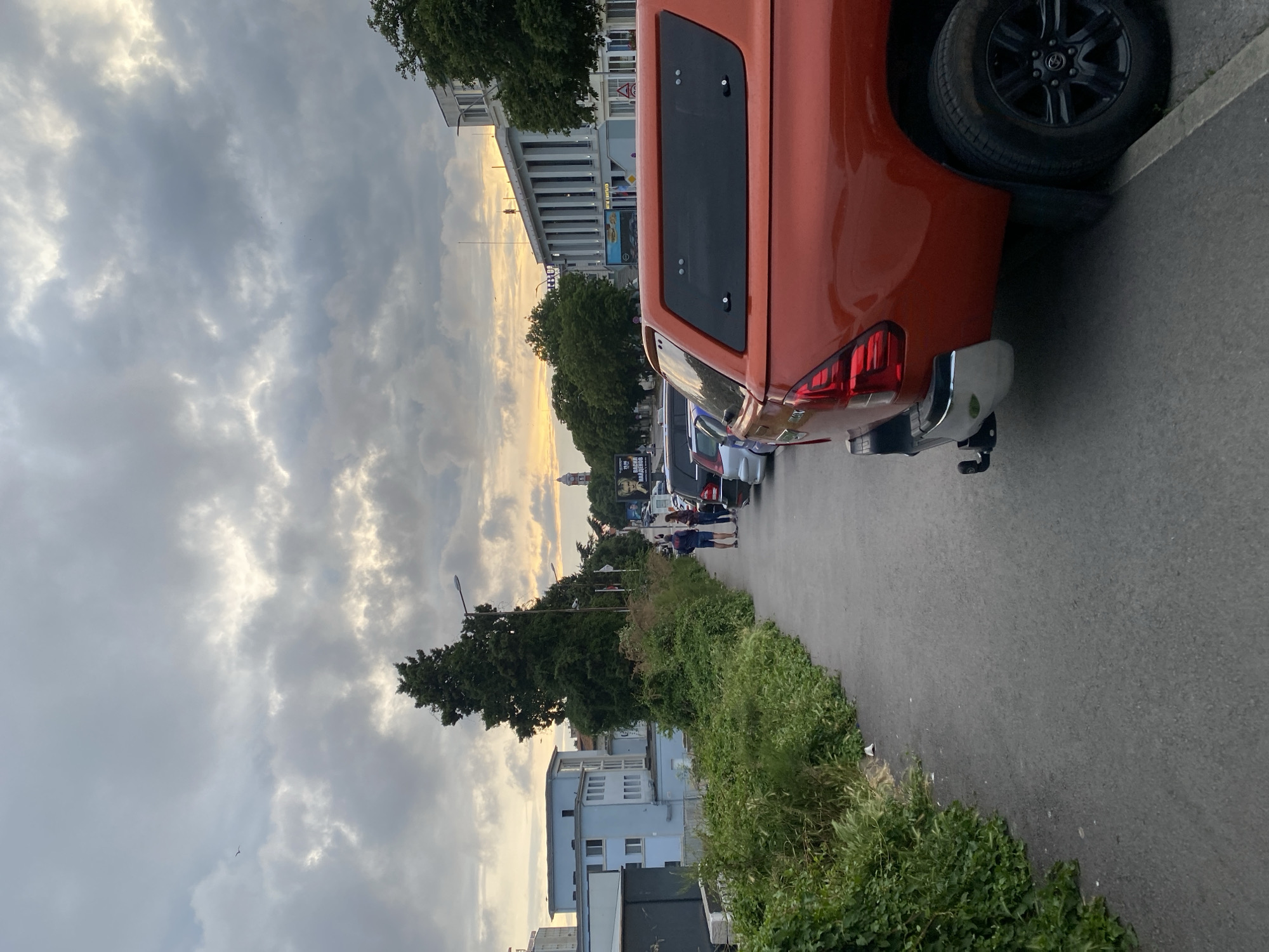 The image displays a street scene in Varna, Bulgaria, captured during what appears to be late afternoon or early evening, characterized by an overcast sky with patches of soft, warm sunlight breaking through. The vantage point is slightly elevated, looking down the length of the street.
In the foreground and prominent on the right is the rear of an orange pickup truck, likely a Toyota Hilux, parked alongside the road. It has a black cargo bed cover, red taillights, and a chrome bumper with a visible tow hitch receiver. Part of its black wheel is visible above the pavement.
The asphalt road stretches into the distance, lined with other parked cars and vibrant green bushes and trees. Several people are visible in the middle ground, standing or walking along the street near the parked vehicles.
On the left side of the street, various buildings line the thoroughfare. Further back, a large, light-colored building with a distinctive facade (possibly columns or vertical slats) is visible, with the faint text "HOTEL & RESTAURANT" discernible on its front. Closer to the foreground on the left is a light blue building with multiple windows and balconies. In the distant background, rising above the buildings and trees, a distinct clock tower stands against the sky. A billboard featuring the partially visible text "MAD DOGS" and other writing can also be seen.
The sky above is dominated by grey, broken clouds, with areas where the sun's soft light penetrates, creating a gentle glow. The overall impression is that of a calm, everyday urban environment.