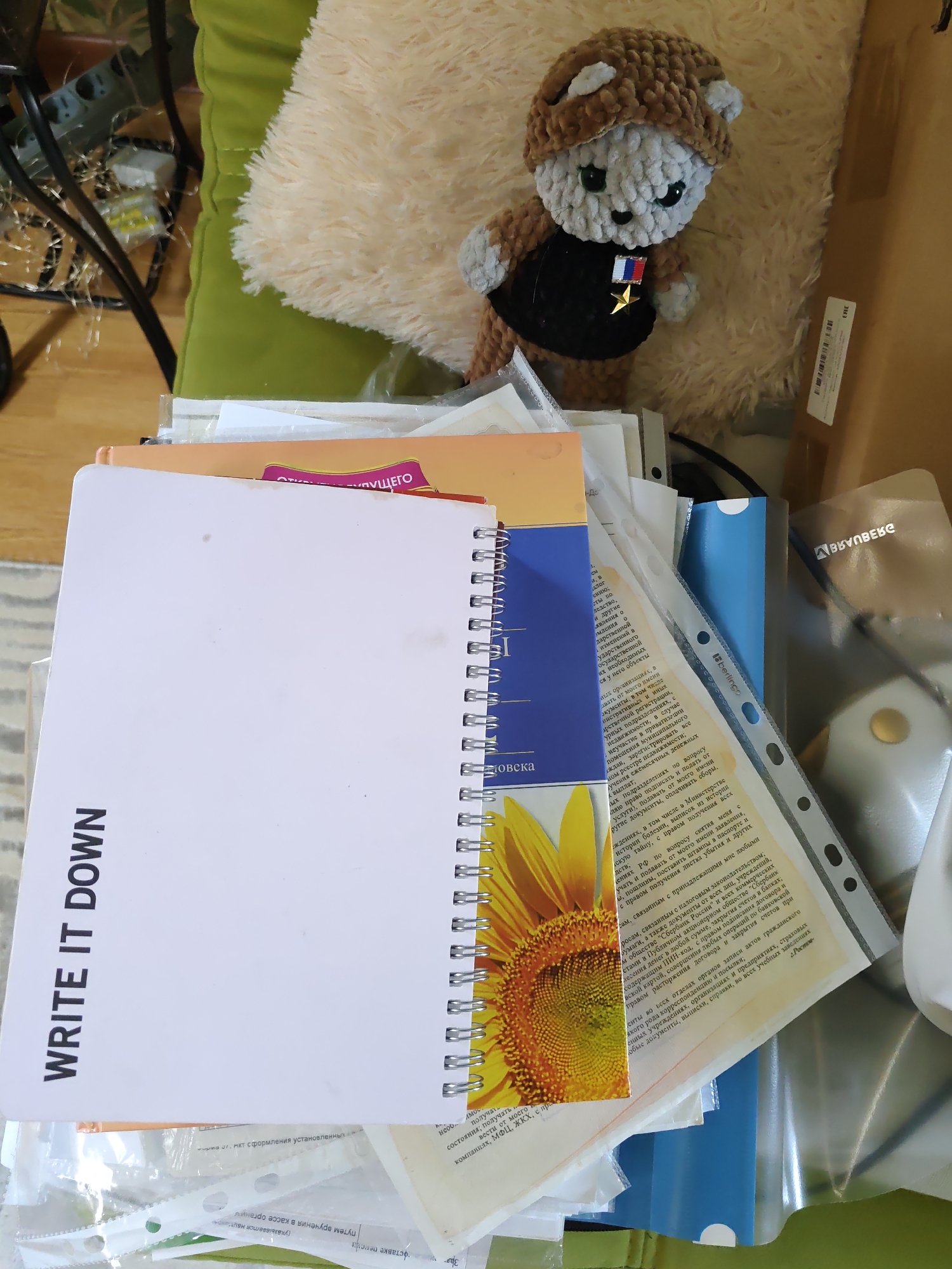 In Rostov-on-Don, Russia, a small, crocheted bear toy wearing a brown hat and a black vest with a medal is positioned on a fluffy, light-colored cushion. The bear has gray fur, green eyes, and appears to be looking slightly to the left. The bear is situated on a wooden surface, near a pile of papers and notebooks. One notebook has the prominent text "WRITE IT DOWN" printed vertically on its cover. Among the papers, there's a printed image of a sunflower. The environment suggests an indoor setting, possibly a desk or table.