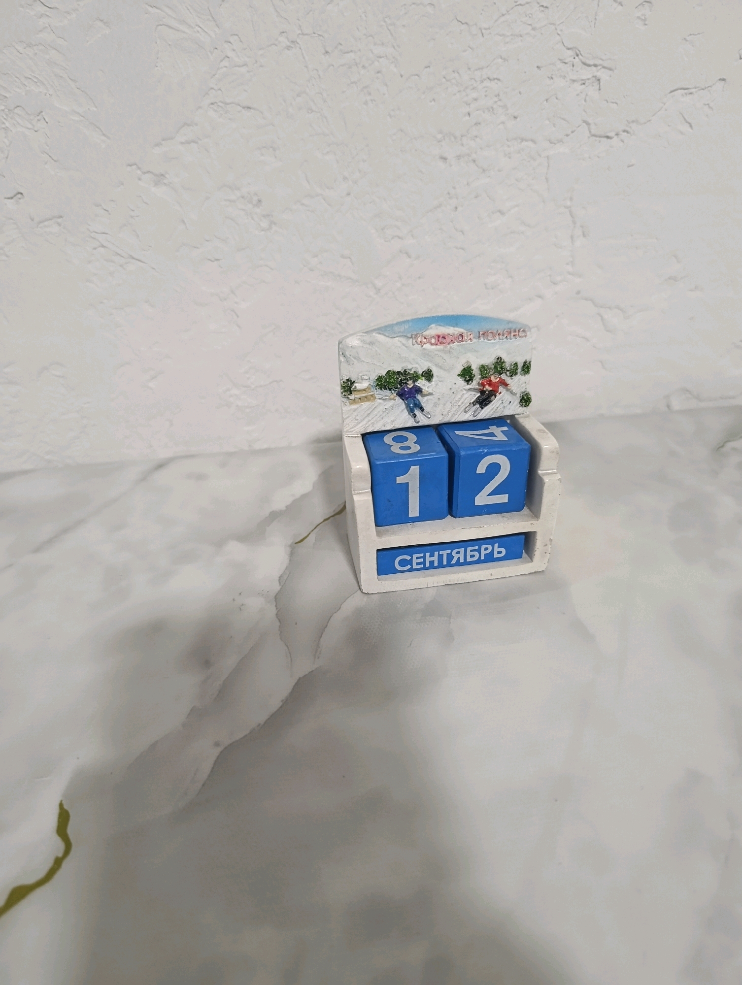 The image shows a close-up of a wooden perpetual calendar. The calendar is white and has a small decorative top featuring a winter mountain scene with two skiers and the text "КРАСНАЯ ПОЛЯНА". The calendar itself consists of three blue blocks, two for the day and one for the month. The day blocks show the numbers "12", with the "1" on the left block and the "2" on the right block. The month block displays the text "СЕНТЯБРЬ" (September). The calendar is placed on a surface with a marbled pattern, and the background is a textured white wall. There is no indication of the city of Арыпжан, Kyrgyzstan in the image.