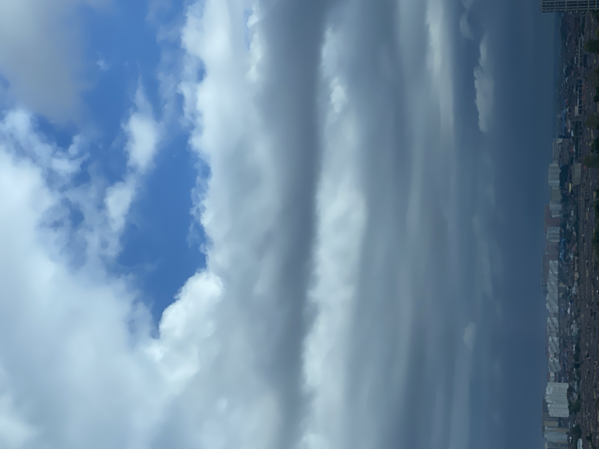 The image depicts an overcast sky over a cityscape, likely Krasnodar, Russia. The sky is filled with various cloud formations, ranging from bright white cumulus clouds in the upper portion to darker, heavier gray clouds lower down, suggesting an impending storm or a shift in weather. Patches of blue sky are visible through thinner areas of the clouds. Below the sky, a dense urban landscape is spread out. Numerous buildings, including apartment complexes and smaller residential structures, are visible. The buildings appear to be of varying heights and architectural styles. Trees and greenery are interspersed among the buildings, indicating a mix of urban development and vegetation. The overall impression is of a cloudy day in a populated area. There are no people visible in the frame. The image is taken from an elevated perspective, looking down on the city.