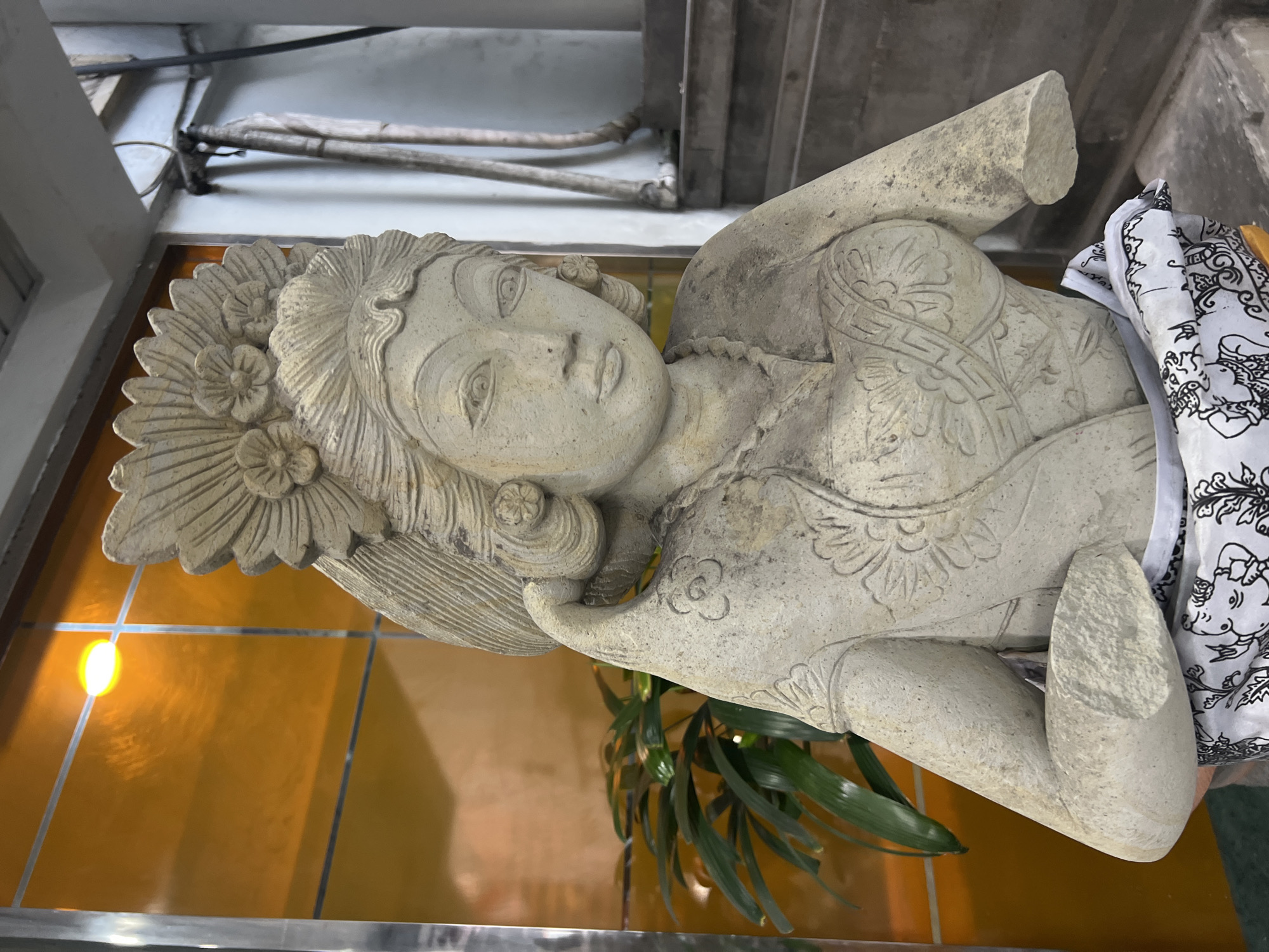 A stone statue of a woman is positioned in front of an orange-tinted window and a lush green plant. The statue, which appears to be ancient or weathered, is dressed in a sarong with a black and white intricate pattern. The woman in the statue has a detailed headdress with flowers and elaborate jewelry. The background shows a staircase and cables, suggesting an indoor or covered outdoor location in Kuta, Indonesia. The statue's arms are missing or broken.
