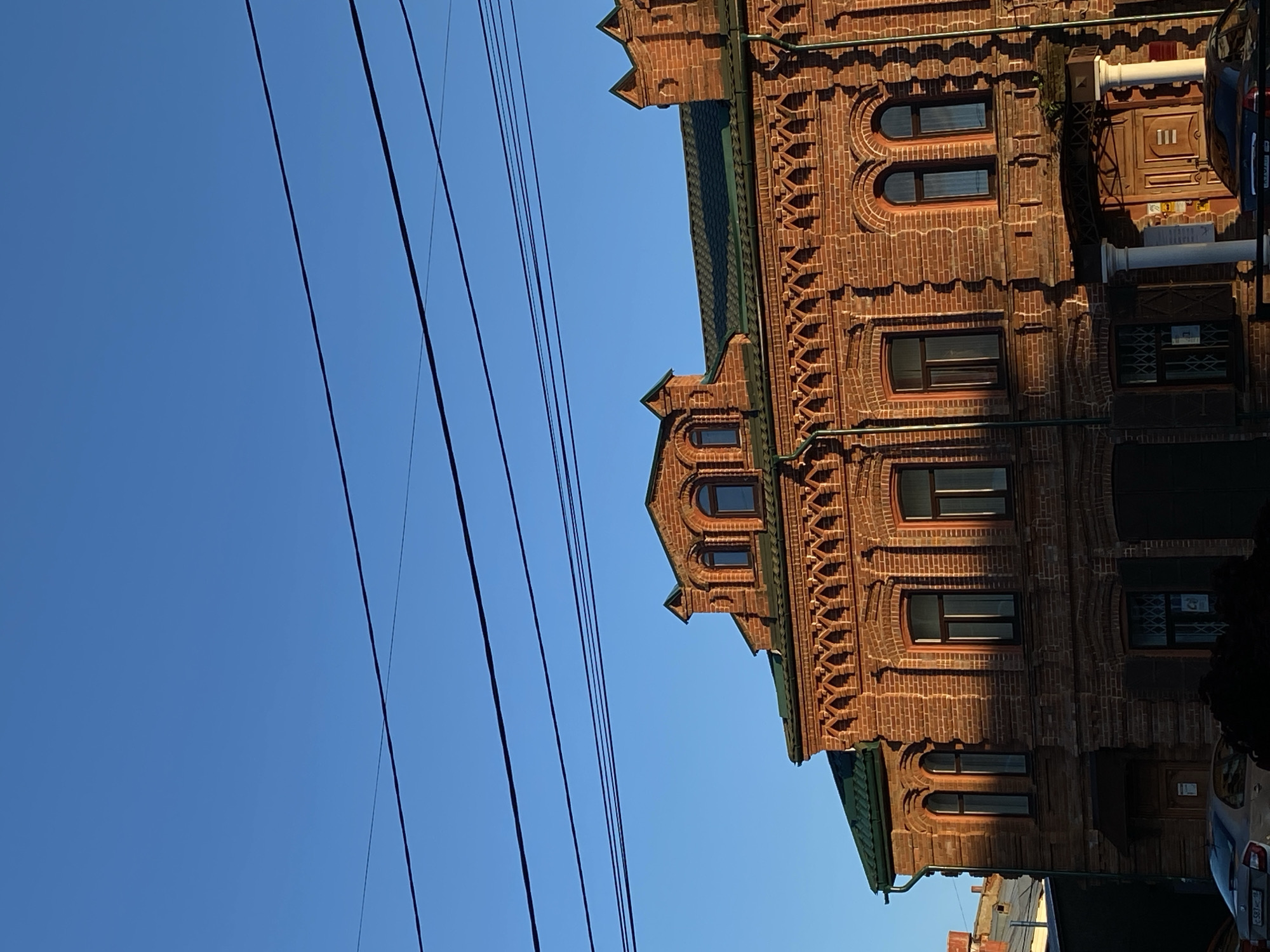The image shows a two-story red brick building with a green roof, situated under a clear blue sky. The building features ornate architectural details, including decorative brickwork above the windows and arched windows on the upper level. Several power lines crisscross the sky in the foreground. The building's facade is partially illuminated by sunlight, casting shadows on the brickwork. The lower level has arched doorways and windows. In the lower left corner, a silver car is partially visible, and a dark object, possibly a bush or low-lying structure, obscures part of the building's base on the right. The overall scene suggests an urban environment in Votkinsk, Russia, likely captured during daylight hours.