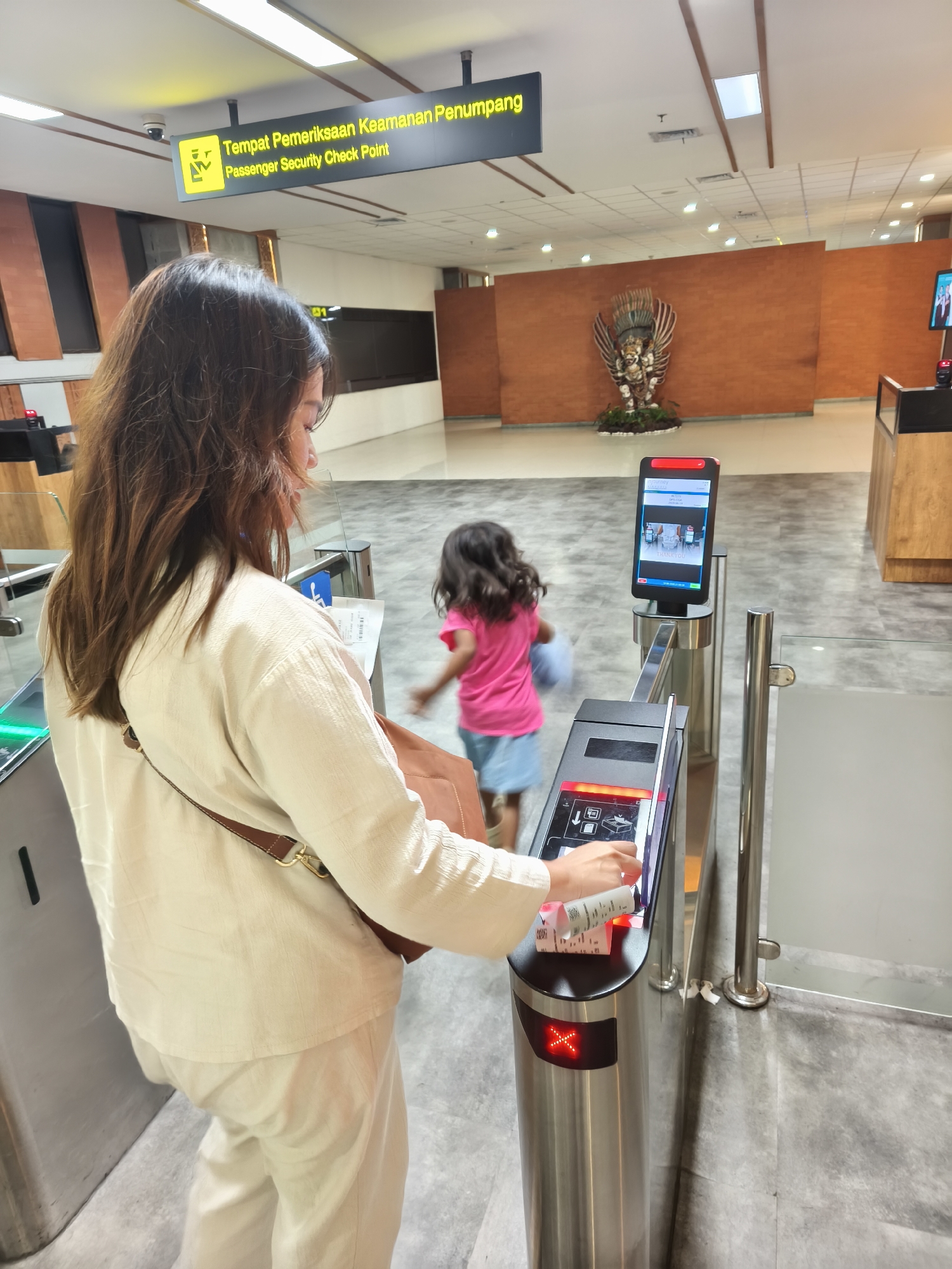 In Kuta, Indonesia, a woman in a cream-colored outfit is holding a ticket or boarding pass as she approaches a security checkpoint at what appears to be an airport. Behind her, a young child in a pink shirt and blue skirt is walking away from the checkpoint. The checkpoint is equipped with a turnstile, a ticket scanner, and a digital display screen showing a "THANK YOU" message. A sign above the checkpoint reads "Tempat Pemeriksaan Keamanan Penumpang Passenger Security Check Point." In the background, a large brick wall features a decorative statue, possibly of a mythological figure, and the overall environment suggests a public space, likely an airport terminal, with modern infrastructure.
