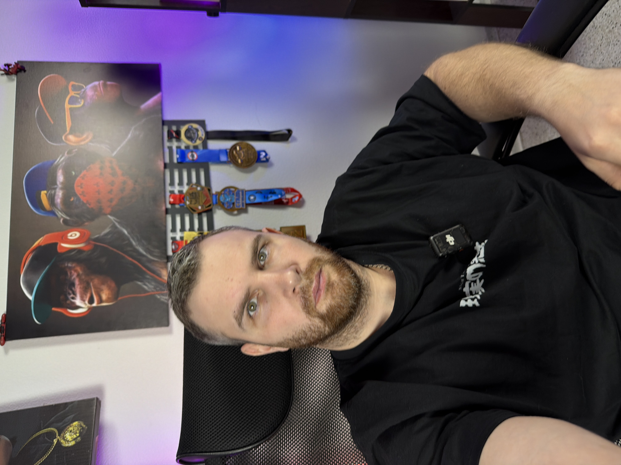 The location is in the city of Unknown, Unknown.
A male individual is seated in a black mesh ergonomic chair, positioned centrally and facing directly towards the camera. He possesses fair skin, short brown hair with visible graying, a trimmed reddish-brown beard and mustache, and green eyes, maintaining a neutral expression. He wears a black short-sleeved t-shirt featuring white text in an unknown script on the chest, and a thin silver-colored chain is visible around his neck. A small, black electronic device, likely a microphone transmitter, is clipped to the right side of his t-shirt. His left arm rests on the chair's armrest.
The background consists of a light-colored wall. To the subject's upper left, a black canvas print depicts a golden medallion on a chain. Centered behind the subject is a larger horizontal canvas print featuring three anthropomorphic monkeys: the leftmost wears a black cap with a teal brim and red headphones, the middle wears a blue cap and a red bandana, and the rightmost wears an orange cap and orange sunglasses. Below this artwork, a black rack displays multiple medals with colorful ribbons, including red, blue, and gold. Ambient purple LED lighting illuminates the wall on the right side of the frame. A dark brown or black shelving unit is partially visible on the far right.